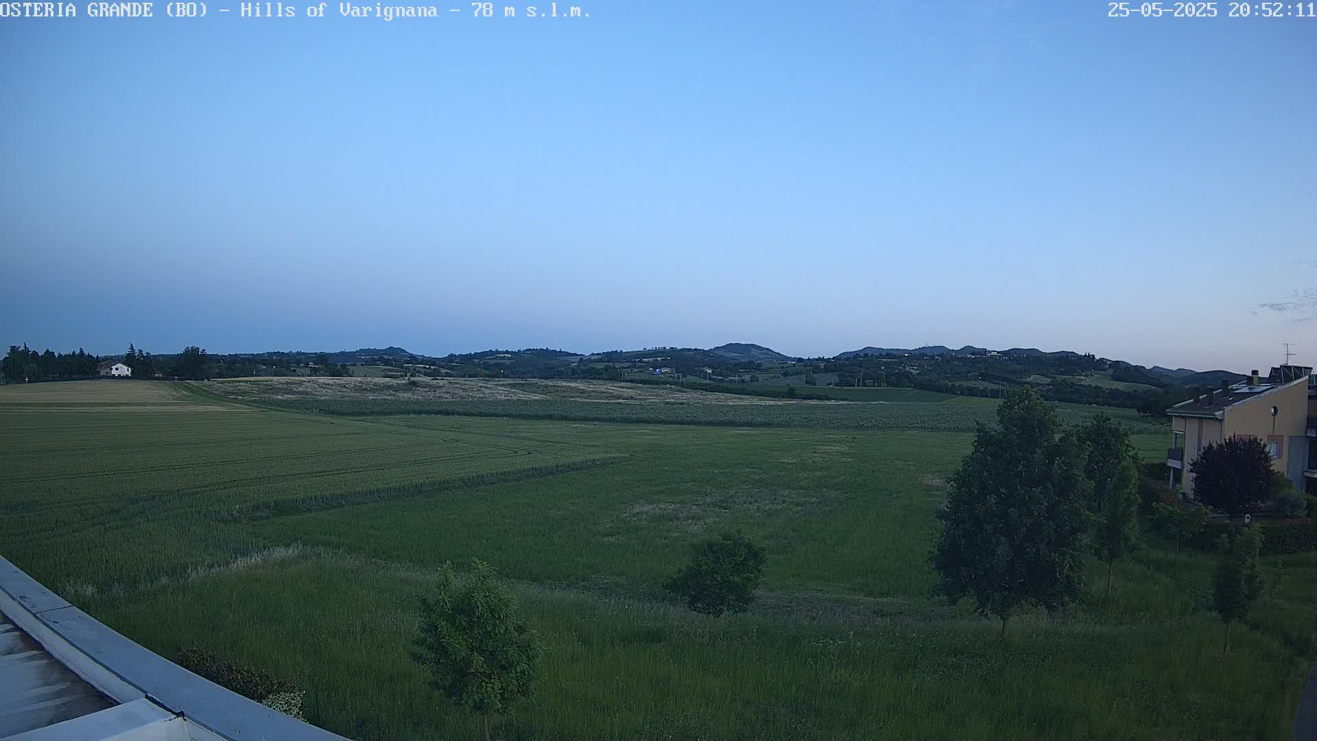 Under a twilight sky, a vast expanse of green fields stretches towards rolling hills in the distance, with a few buildings visible near the bottom right.