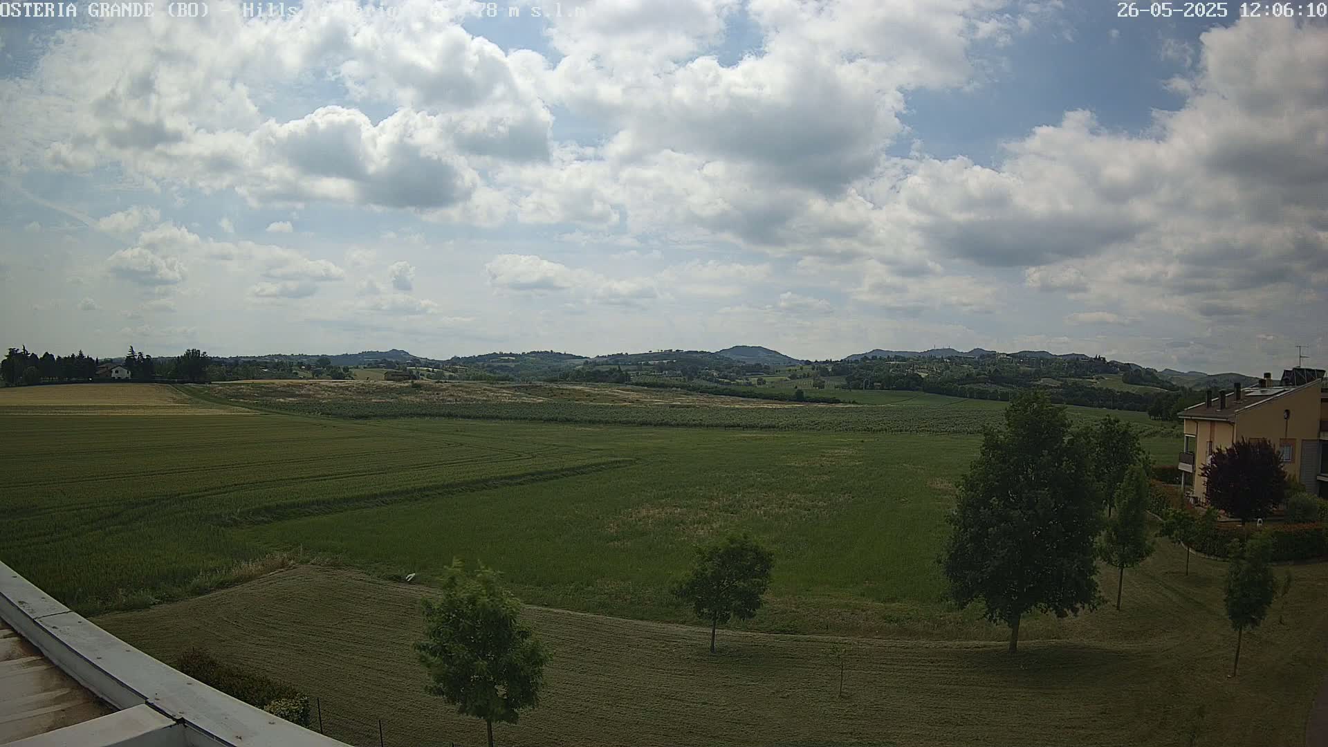 A partly cloudy sky overlooks a vast expanse of green fields and distant hills, with a few trees and a building visible in the foreground.