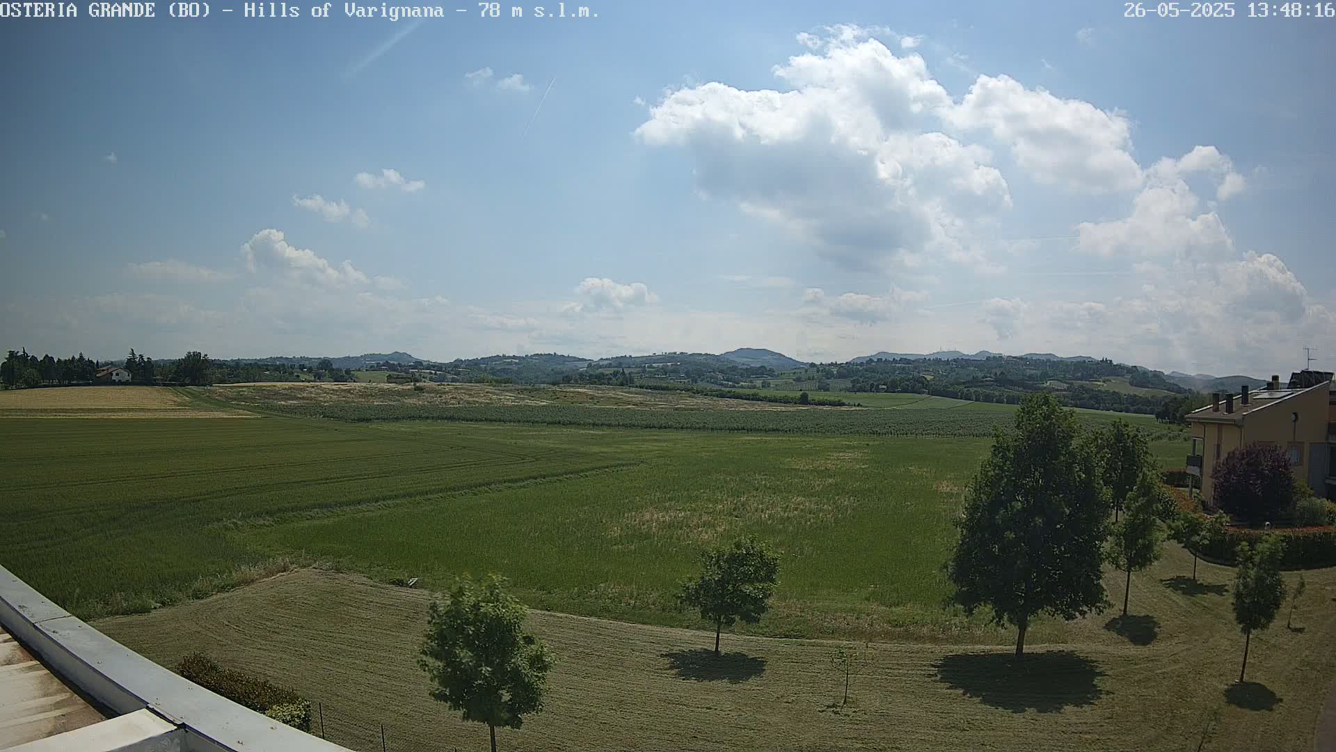 A partly cloudy day reveals a sprawling green field, dotted with trees, extending towards rolling hills in the distance.