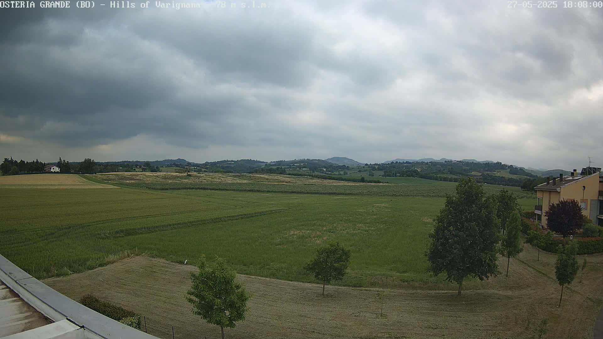 Under a cloudy sky, a wide expanse of green fields stretches towards rolling hills in the distance, with a few trees and a building visible in the foreground.