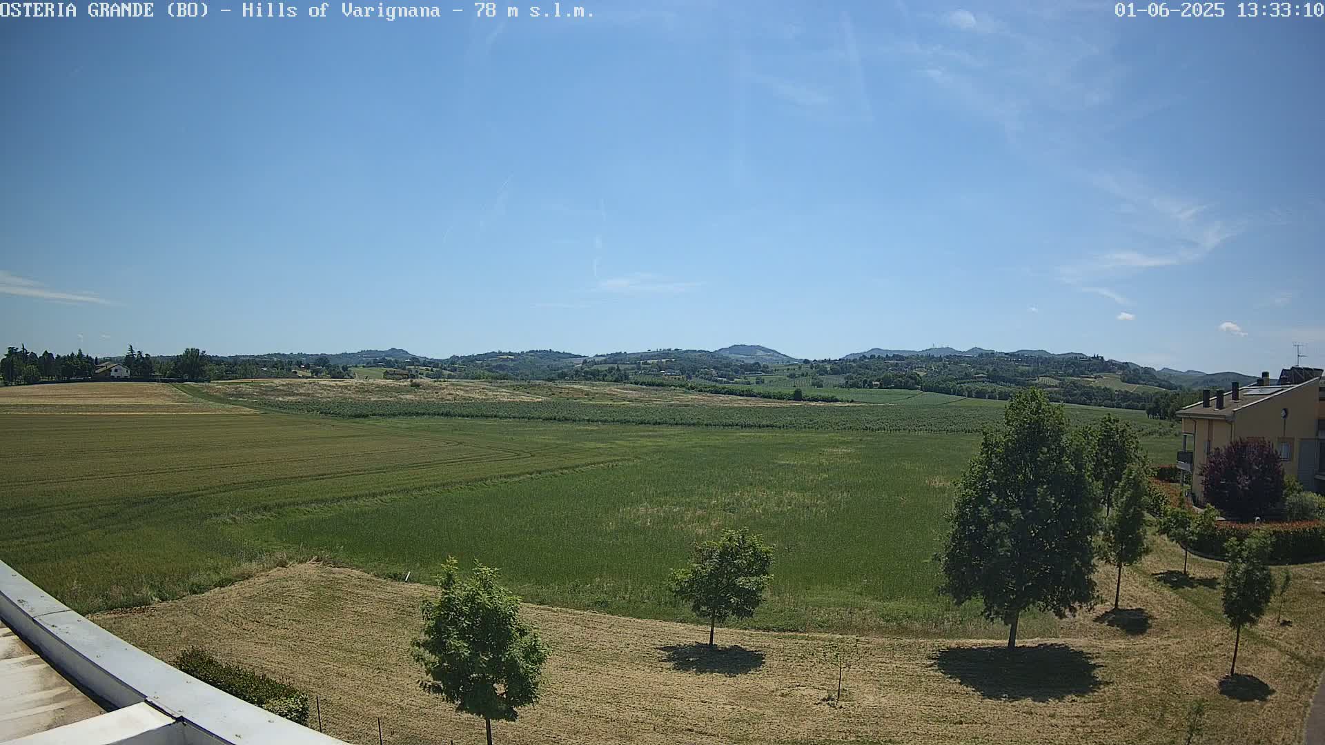 Under a clear blue sky, a sprawling green field extends to rolling hills in the distance, with a few trees and a building visible near the foreground.