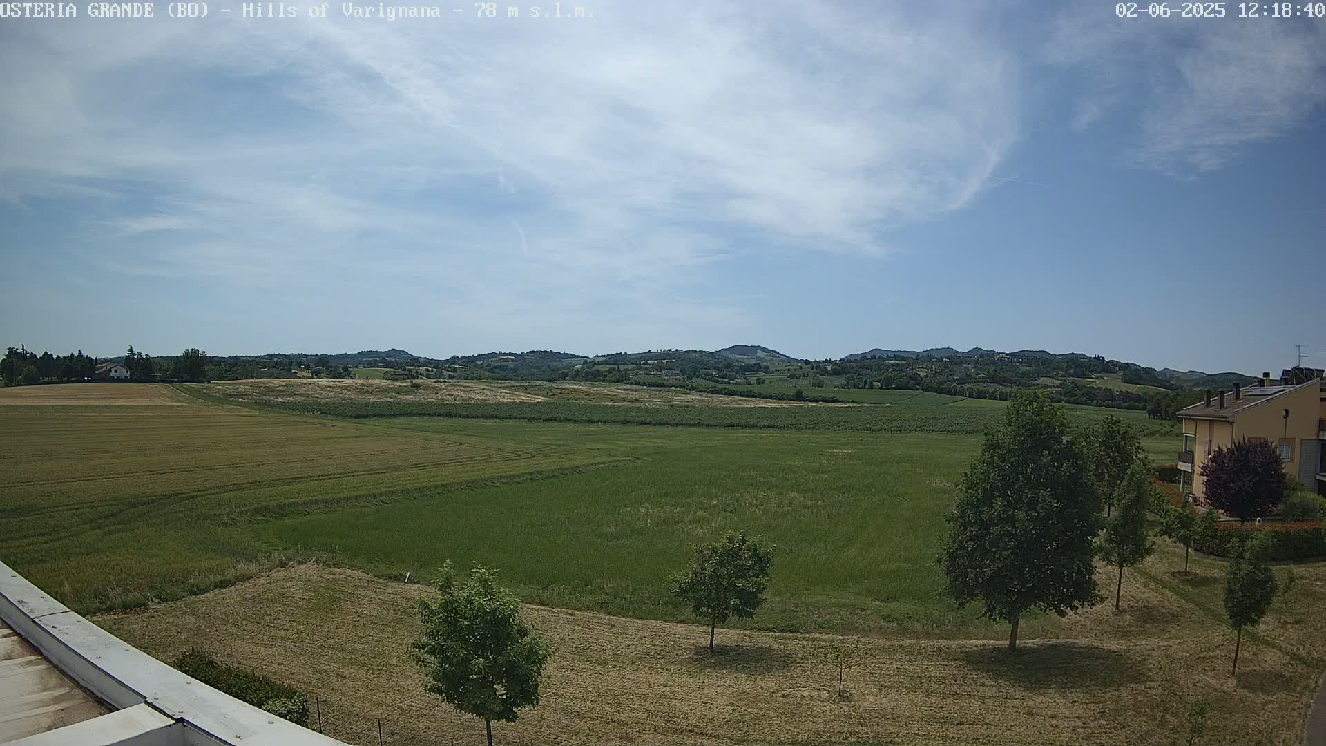 A partly cloudy sky looks over expansive green and brown fields, a few trees, and a building in the distance.