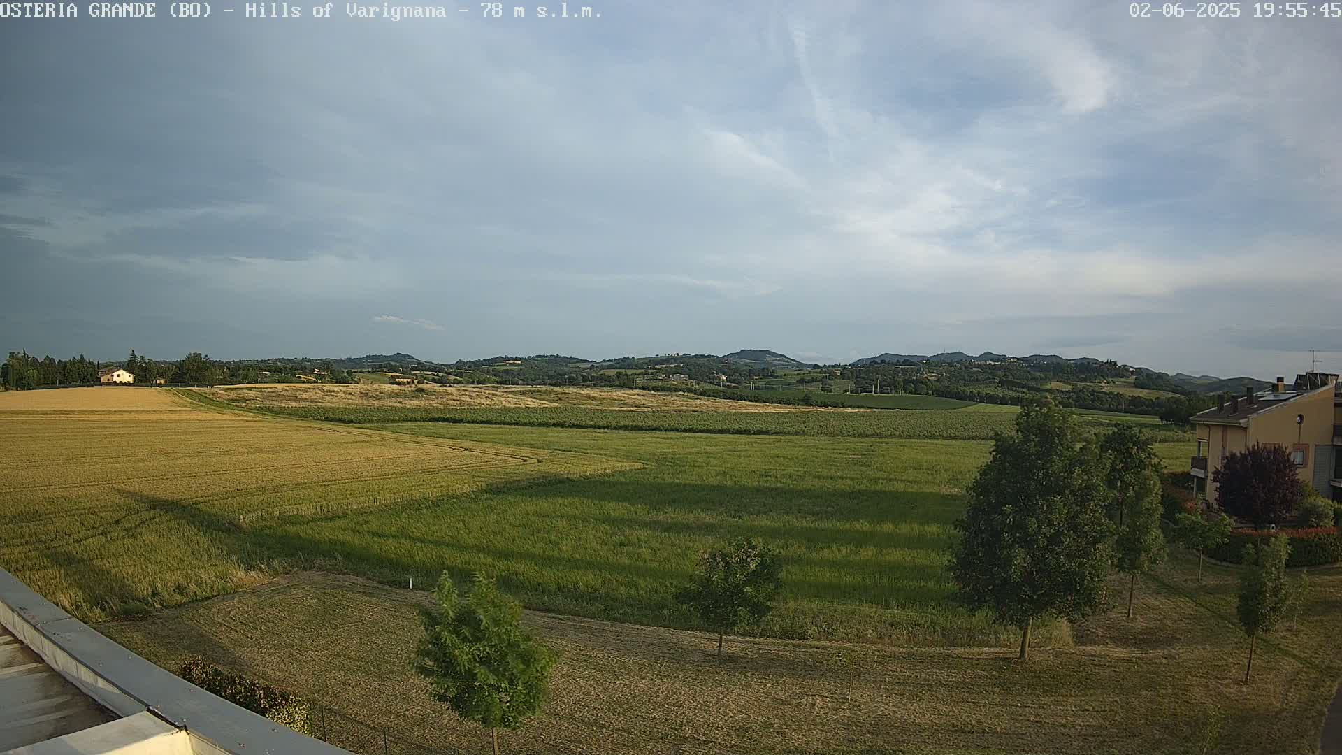 A partly cloudy sky overlooks expansive fields of golden and green crops, a few buildings, and scattered trees under a mostly cloudy sky.