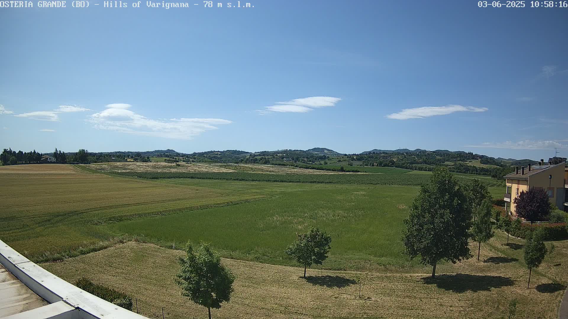 A partly sunny day reveals a vast expanse of farmland with rolling green hills in the distance, a few scattered trees, and a residential building at the lower right.