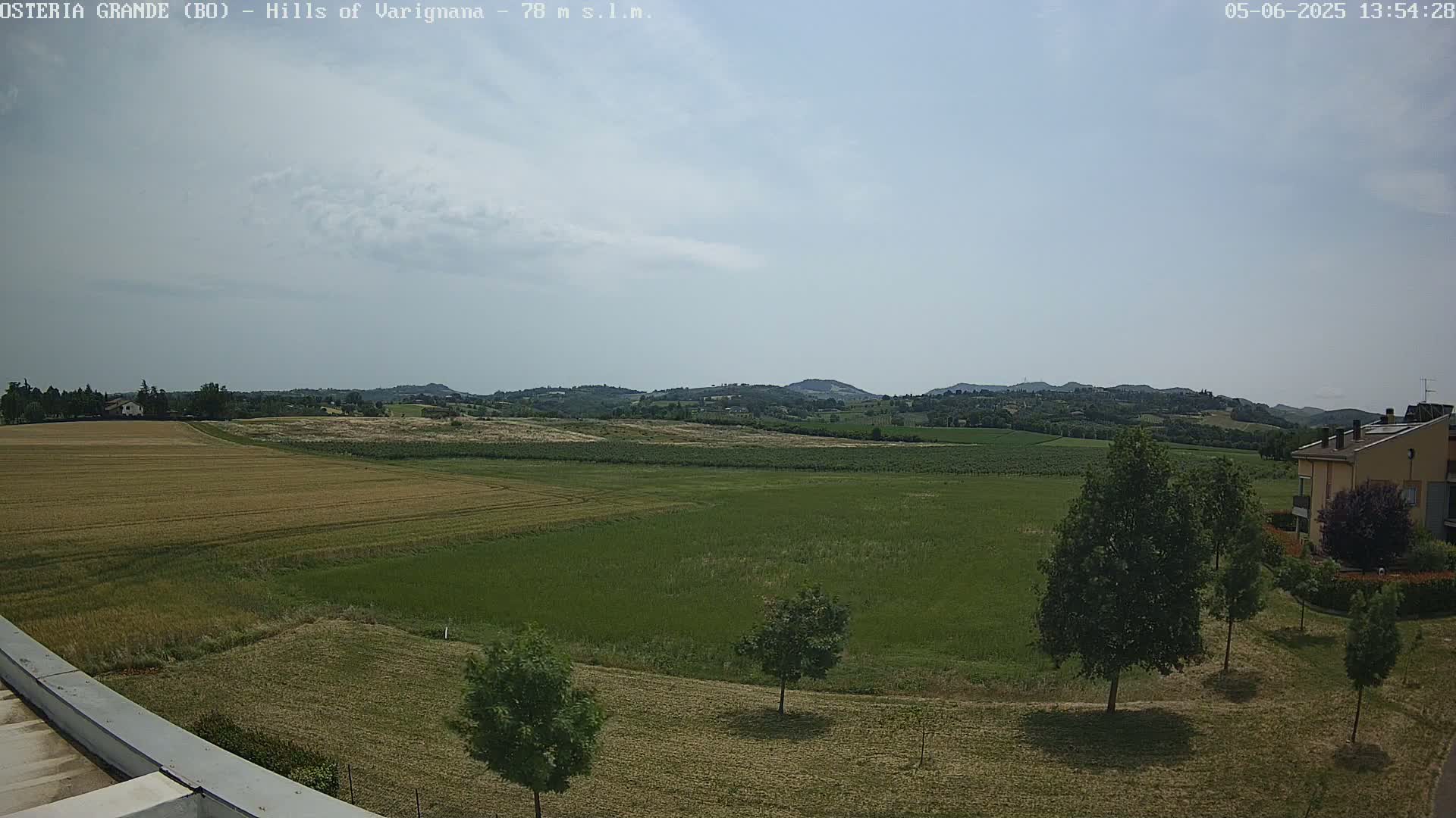A partly cloudy day reveals a wide expanse of farmland, dotted with trees, leading to rolling hills in the distance.