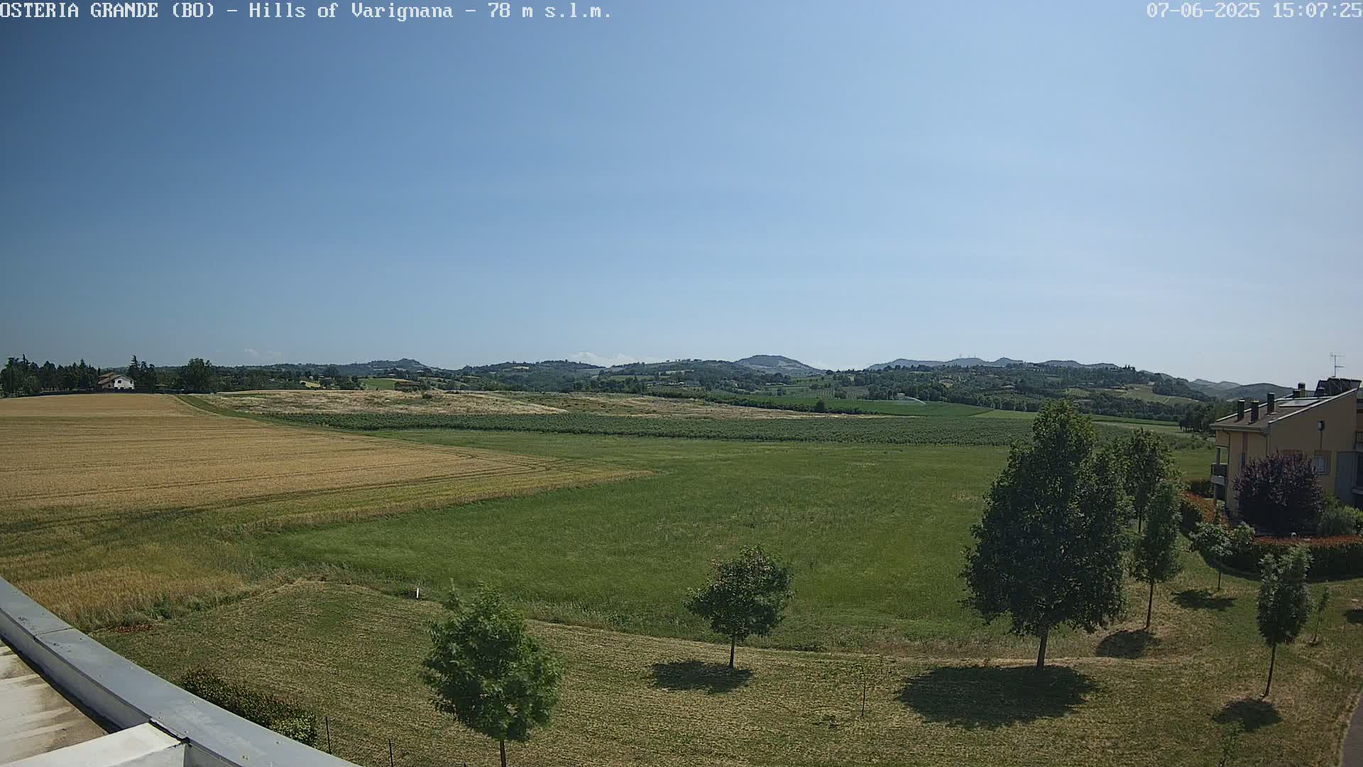 A wide, sunny outdoor view shows a flat expanse of green and golden fields, bordered by trees and a few buildings, with rolling hills in the distance under a clear blue sky.