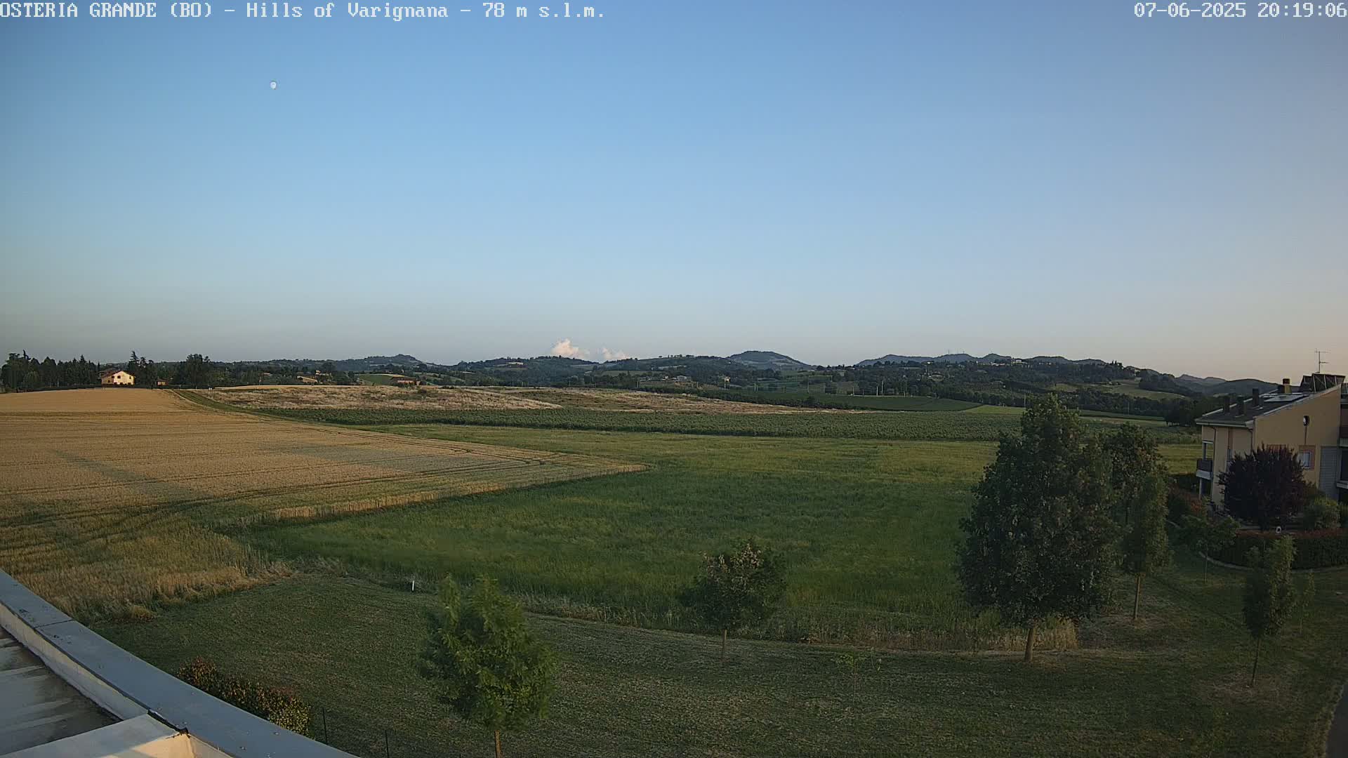 A wide, flat expanse of farmland with golden and green fields stretches toward rolling hills under a clear, light-blue sky.