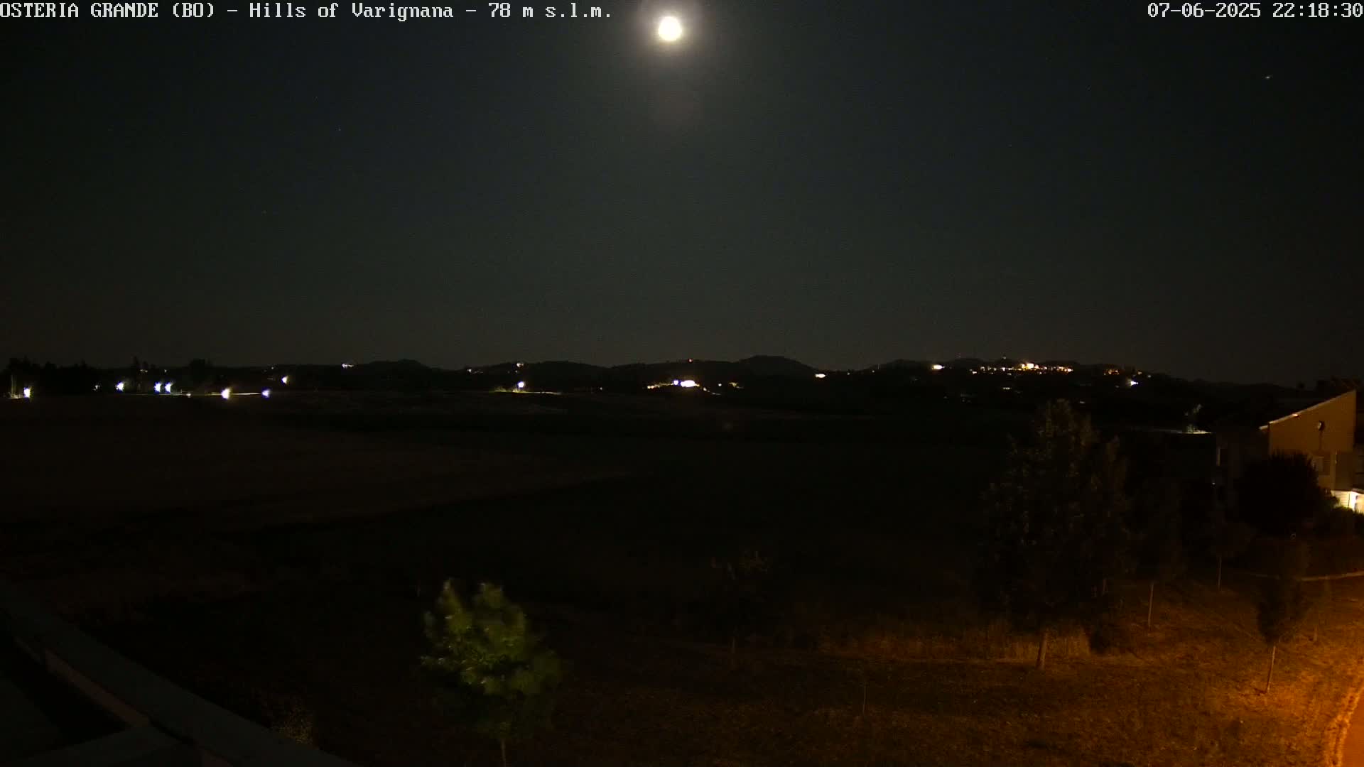 A nighttime view of dark hills with a few distant lights under a bright moon and clear sky.