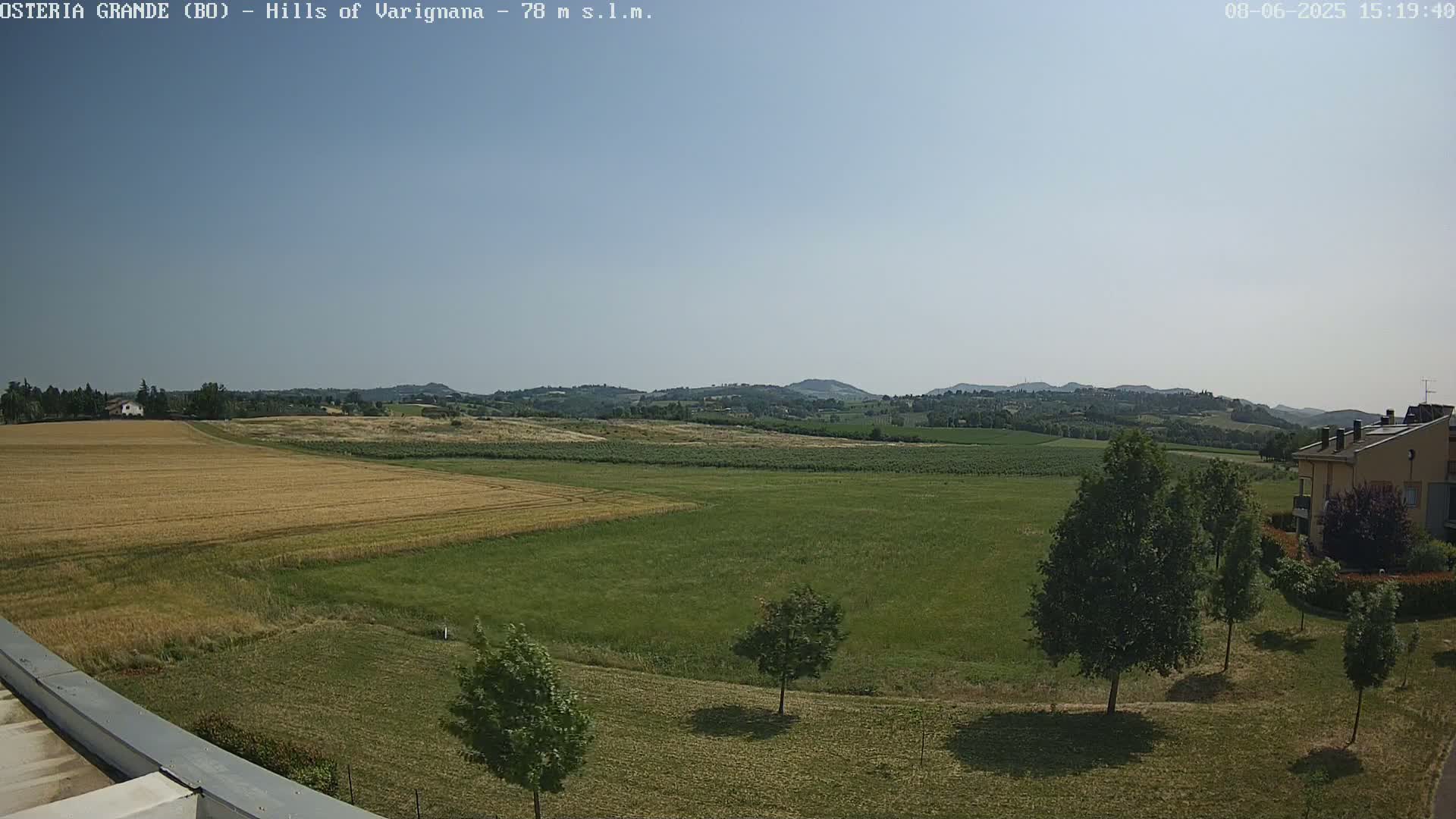 A mostly sunny view of a sprawling green and golden field, dotted with trees, extends to rolling hills in the distance.