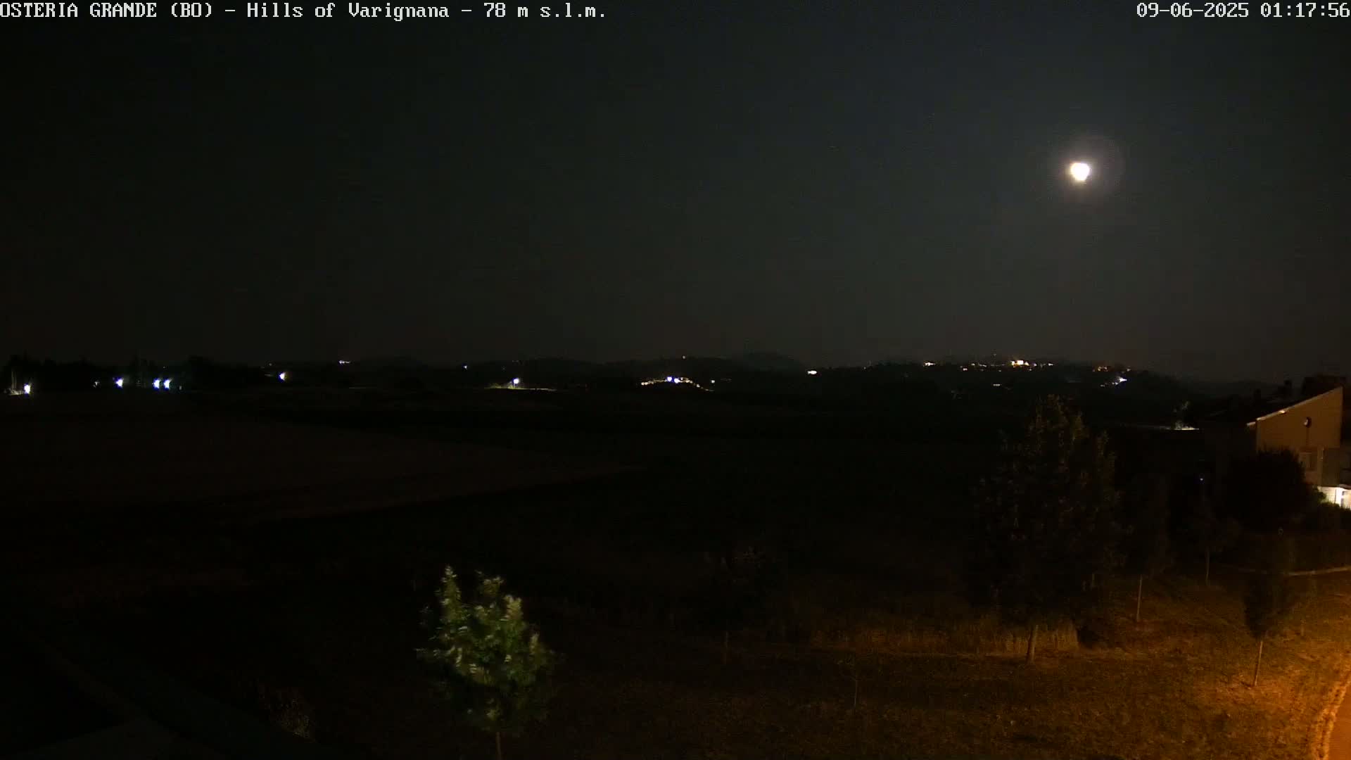 A nighttime view shows a bright moon in a dark sky above a landscape of dark hills with sparsely scattered lights, and some vegetation in the foreground.