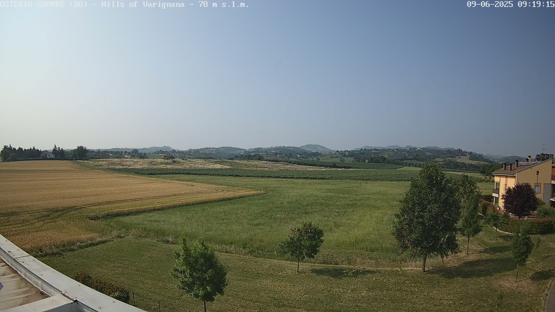 A clear, sunny day reveals a vast landscape of golden wheat fields, green meadows, and distant rolling hills, with a few buildings and trees visible in the foreground.