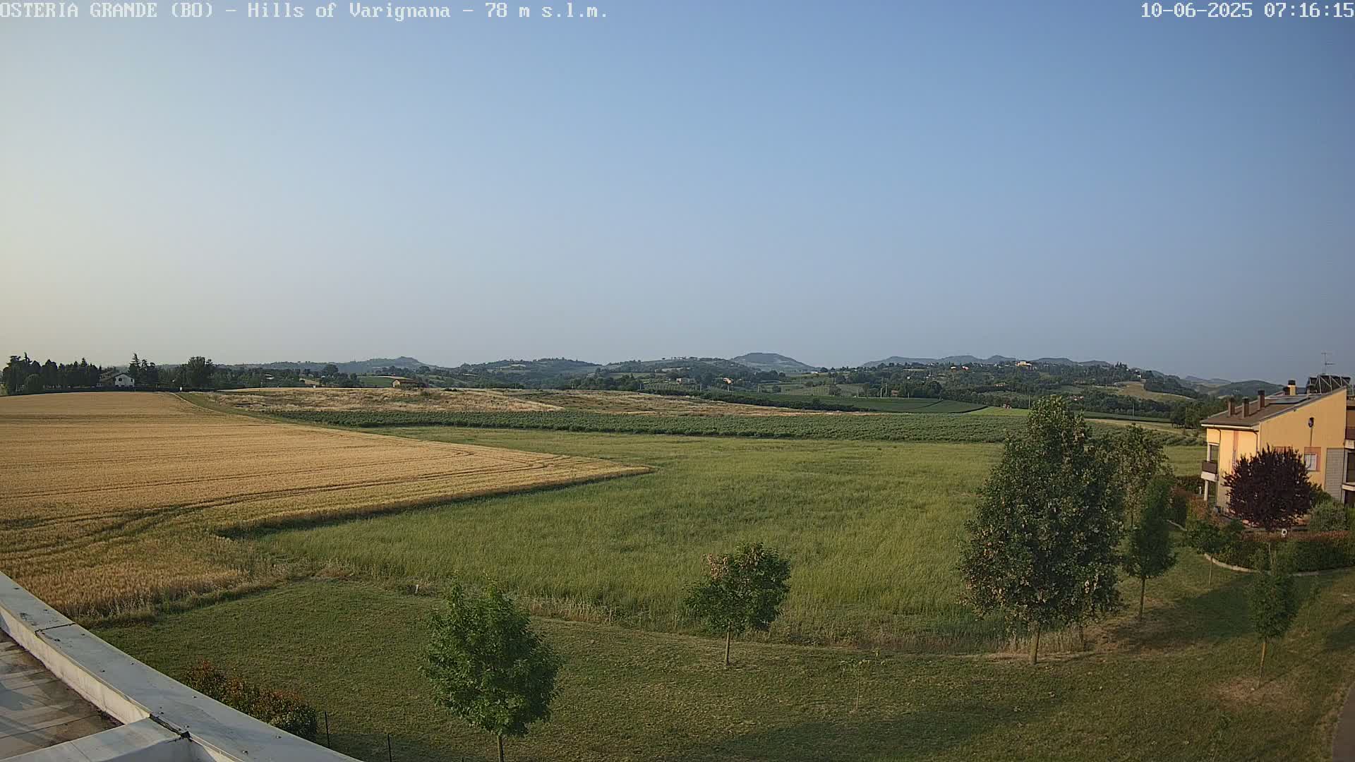 A wide, expansive view of a rural landscape under a clear, light-blue sky shows fields of golden wheat and green grass, rolling hills in the distance, and a few buildings near the bottom right.