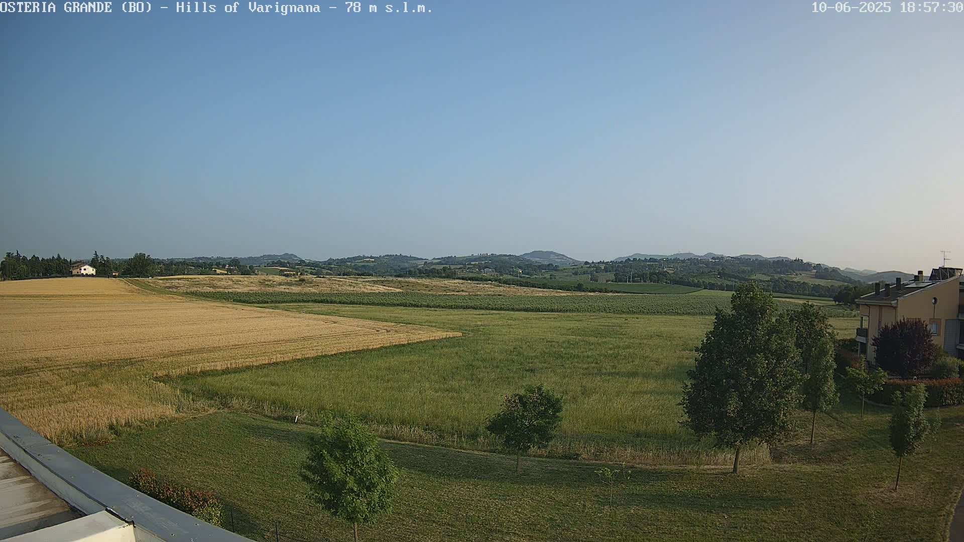A wide, expansive view of rural farmland under a clear, light blue sky shows golden wheat fields and green pastures, with a few trees and buildings visible in the distance.