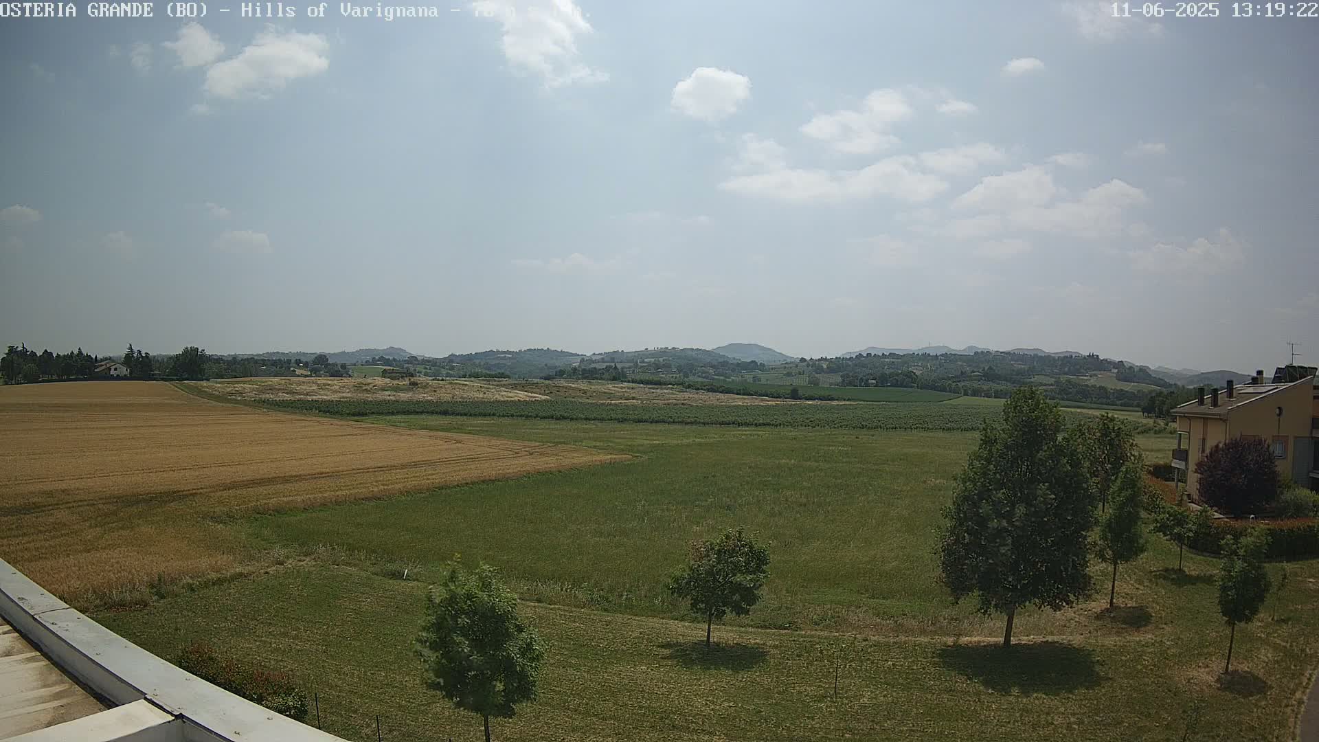 A partly cloudy day reveals a wide expanse of farmland with golden wheat fields and green pastures, rolling hills in the distance, and a few trees and buildings near the bottom of the frame.
