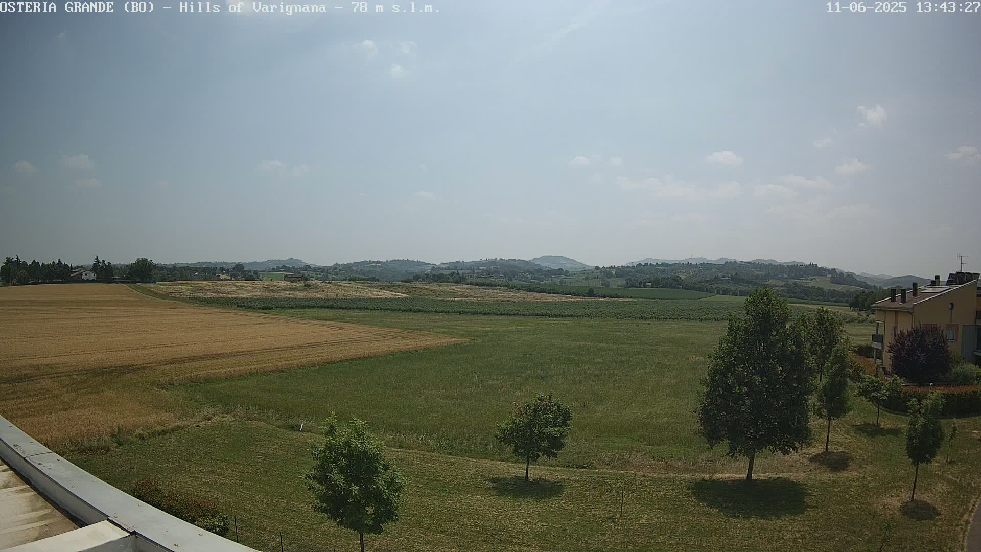 A mostly sunny view of a rural landscape with fields of golden-brown and green grass, some trees, and a building in the distance under a partly cloudy sky.