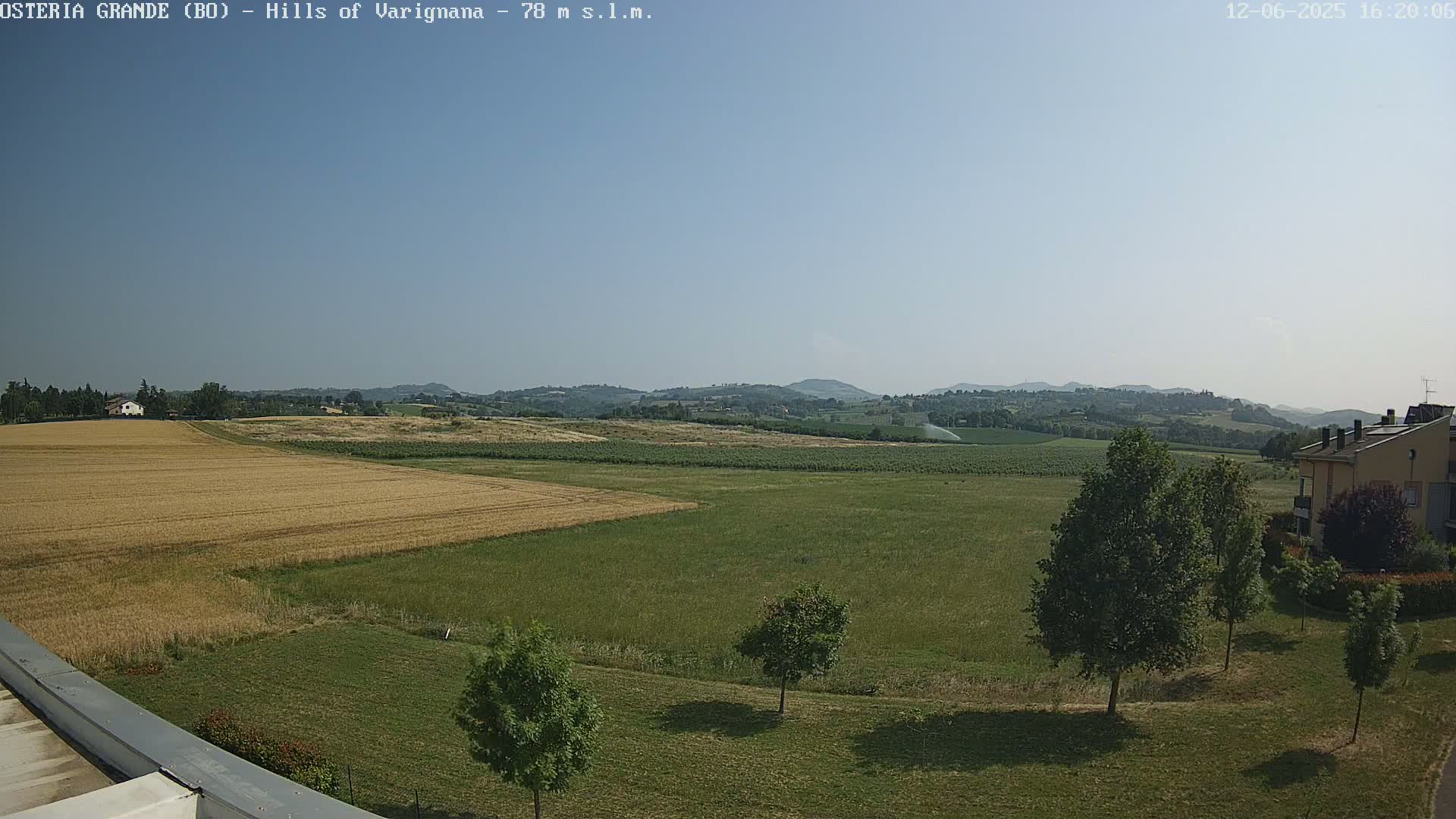 A mostly sunny view of rolling hills and farmland, with a few trees and buildings in the foreground.