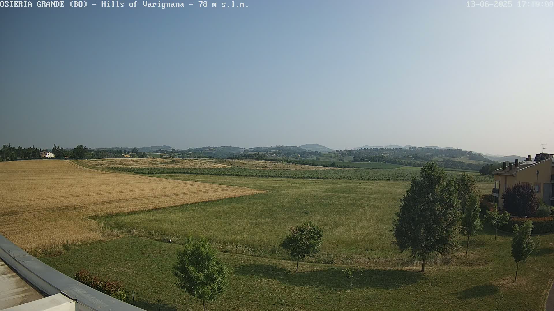 A mostly sunny view of rolling hills and farmlands, featuring a golden wheat field and a building in the lower right corner.