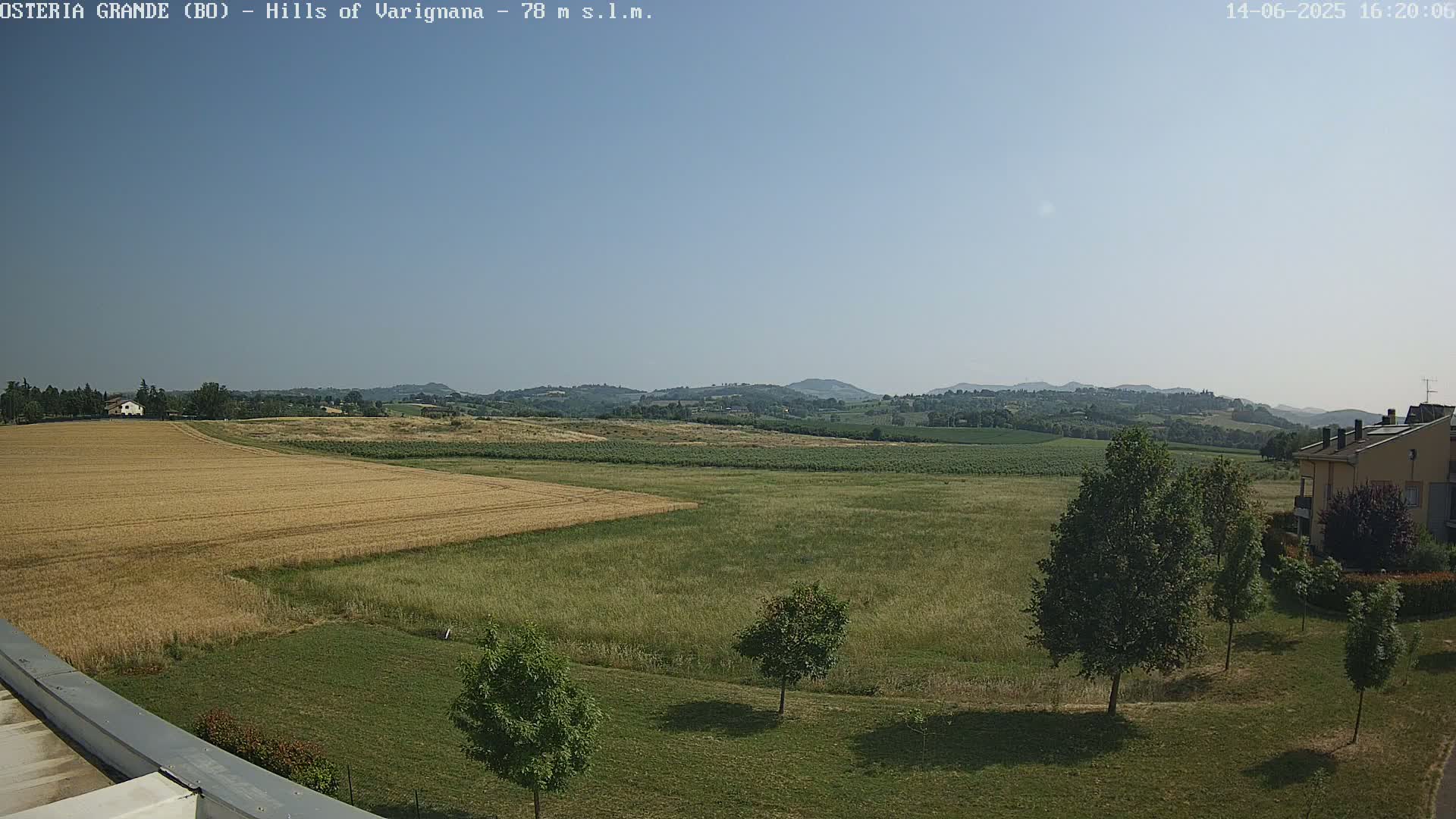 A sunny, clear day reveals a landscape of golden-brown and green fields, with a few trees and a distant line of hills.