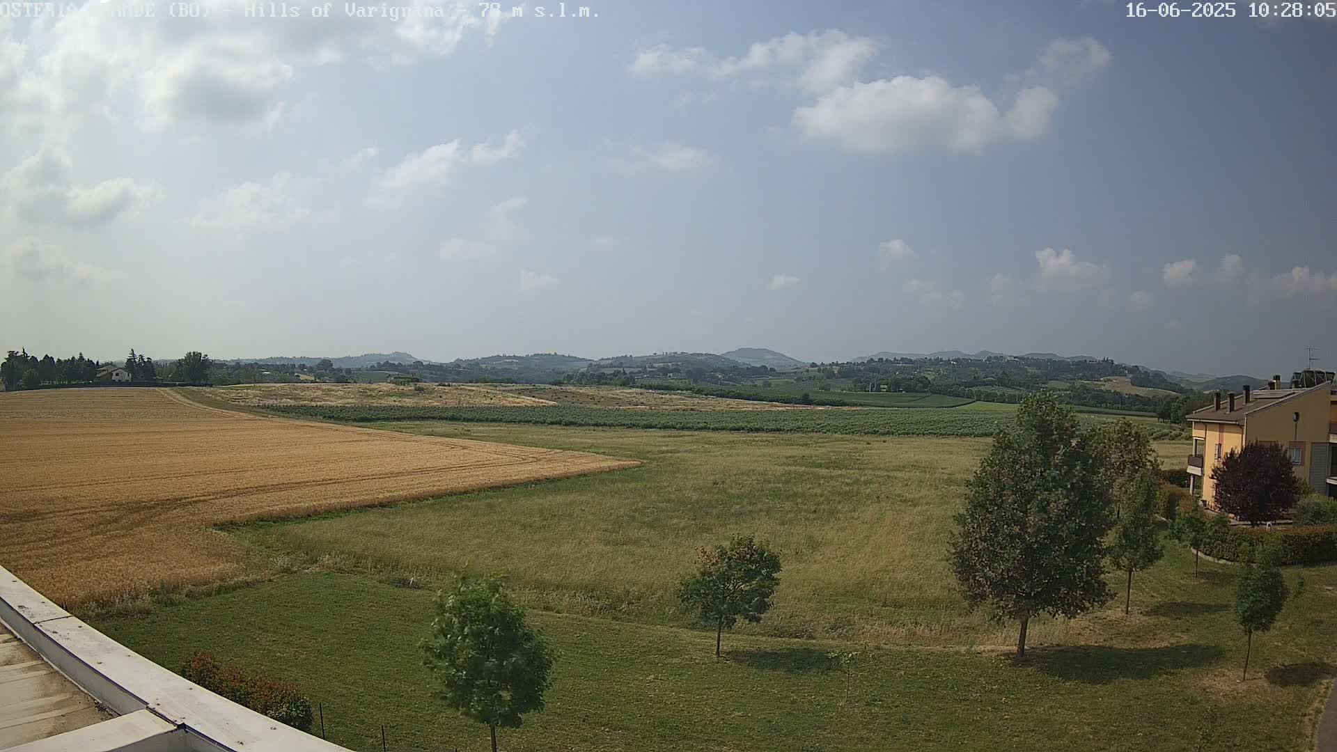 A partly cloudy sky overlooks a landscape of golden wheat fields, green pastures, a few trees, and a yellow building nestled amongst rolling hills in the distance.