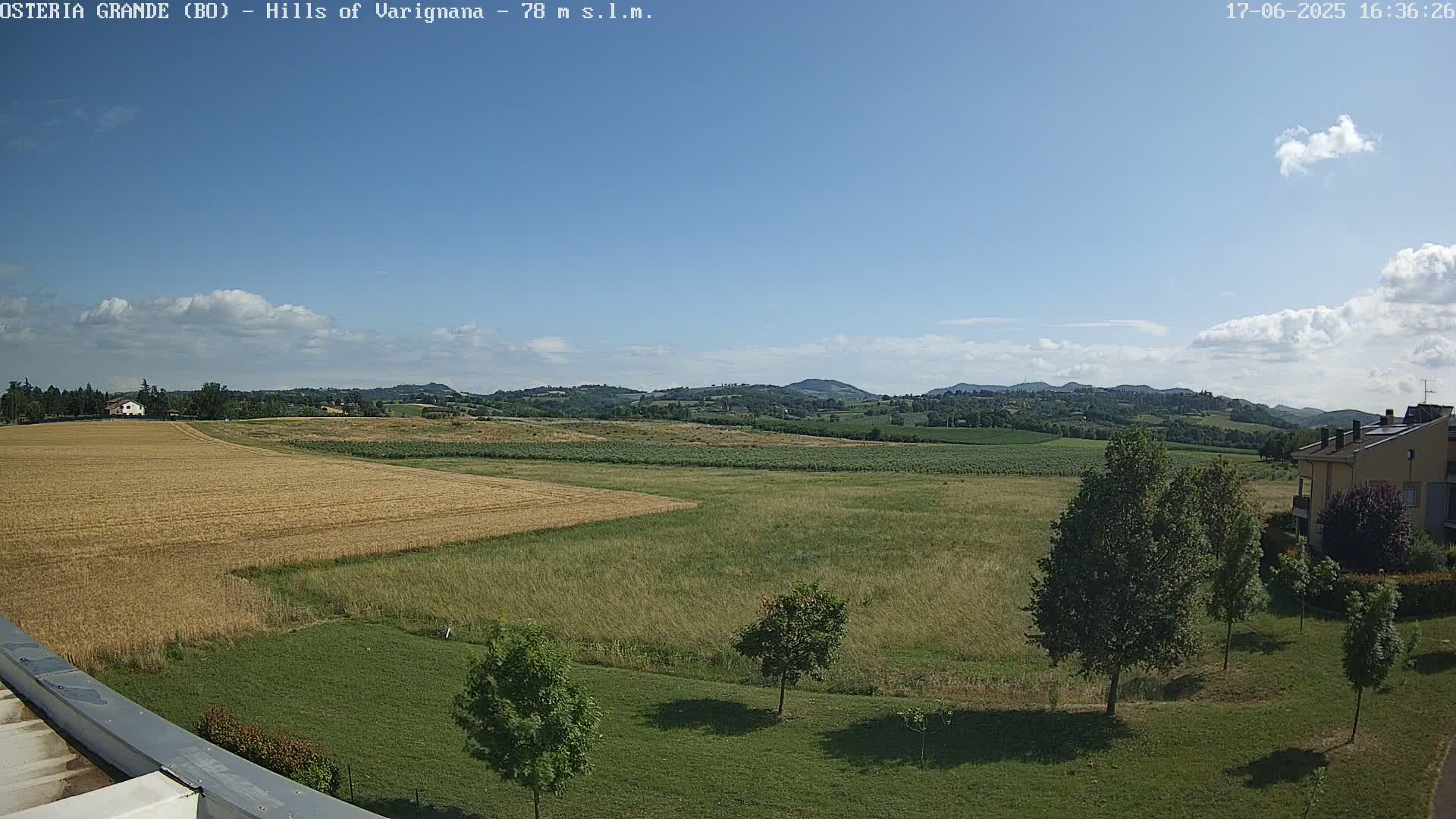 A partly cloudy blue sky overlooks a landscape of golden and green fields, a few buildings, and some trees, with rolling hills in the distance.
