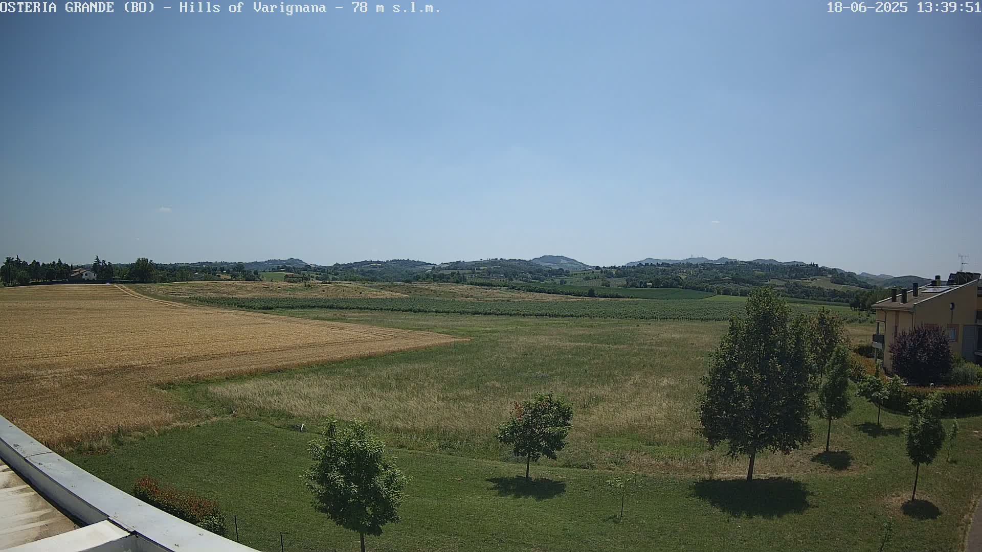 A sunny day reveals a vast expanse of golden wheat fields and verdant meadows, stretching towards a line of rolling hills under a clear blue sky.