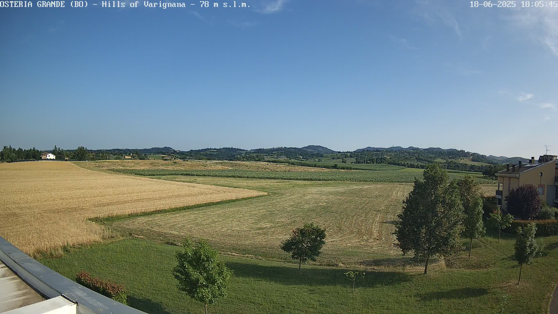 A mostly sunny view of rolling hills and agricultural fields, with a few trees and a building in the foreground.
