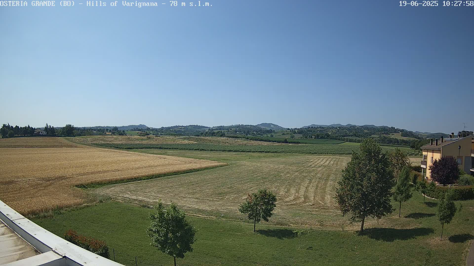 Under a clear blue sky, a rural landscape unfolds with fields of harvested crops, green pastures, and a few trees near a building, all nestled against a backdrop of rolling hills.