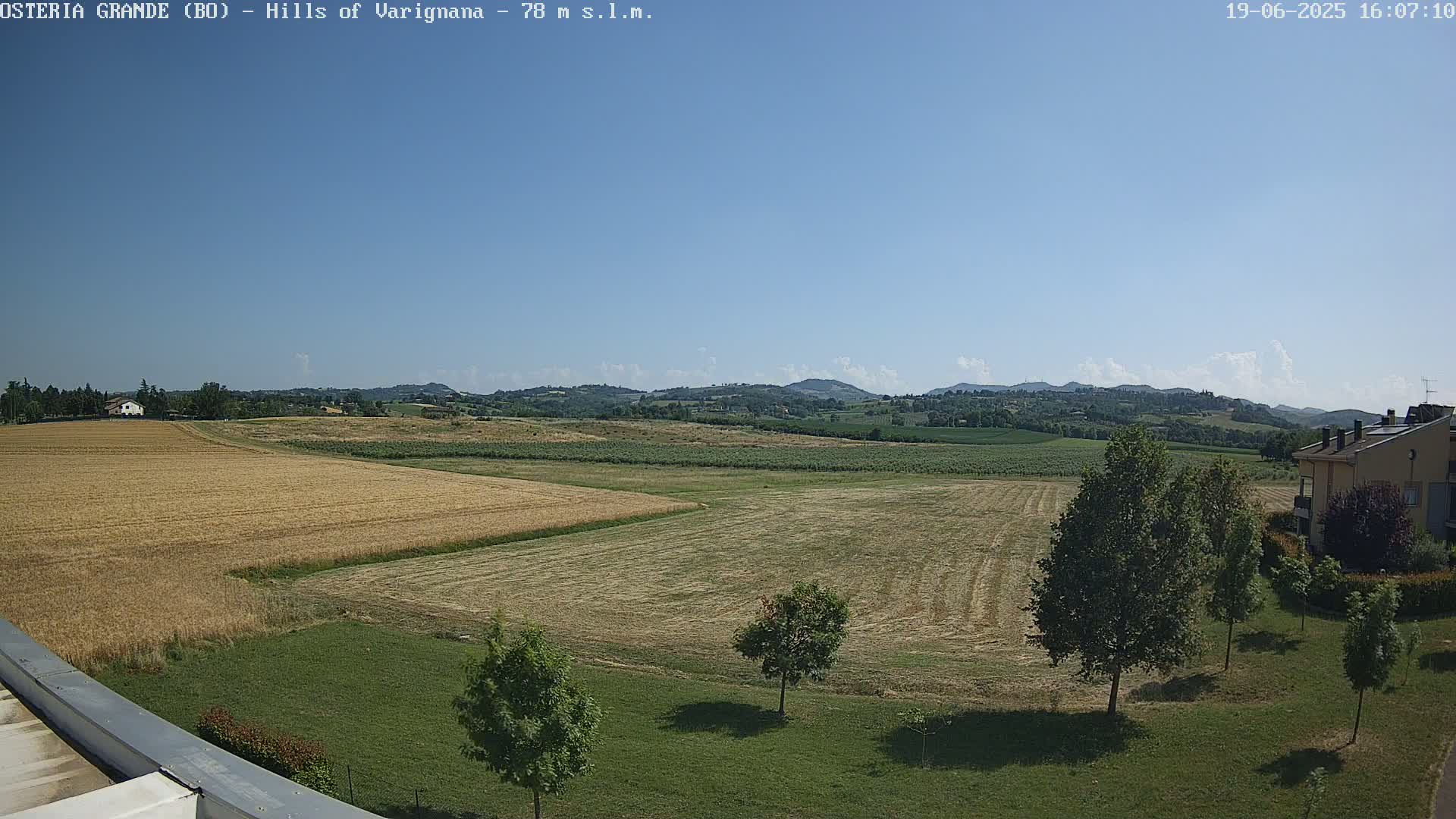 A sunny, clear day reveals a rural landscape of harvested fields, green lawns, and distant rolling hills, with a few buildings visible near the bottom right.