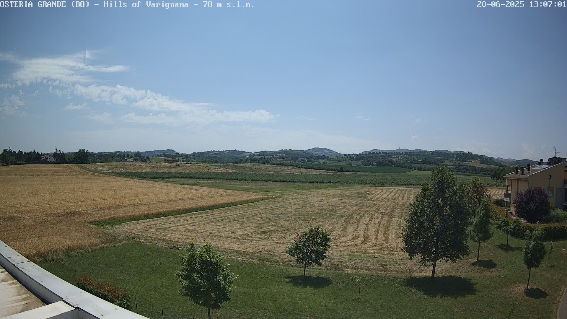 A mostly sunny day reveals a wide expanse of harvested fields and rolling hills in the distance, with a few trees and a building visible in the foreground.