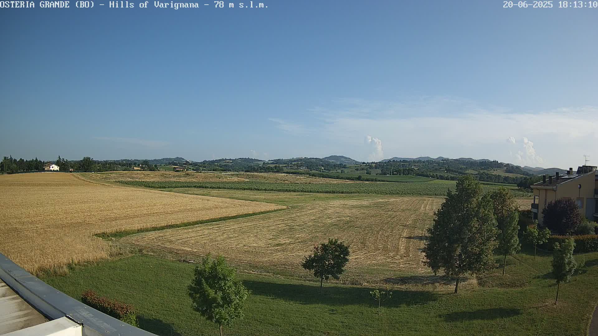 A mostly sunny day reveals a wide expanse of harvested fields and verdant hills in the distance, with a few trees and buildings visible closer to the foreground.