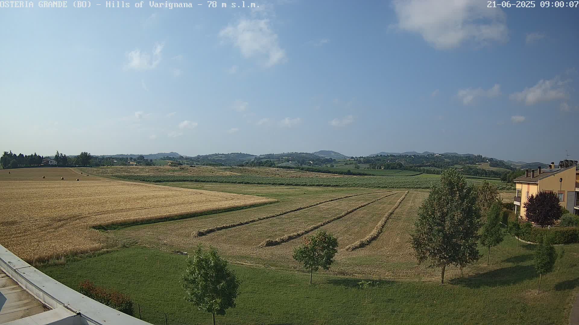 A partly cloudy sky overlooks a rural landscape of harvested and unharvested fields, with rows of hay and a building in the background.