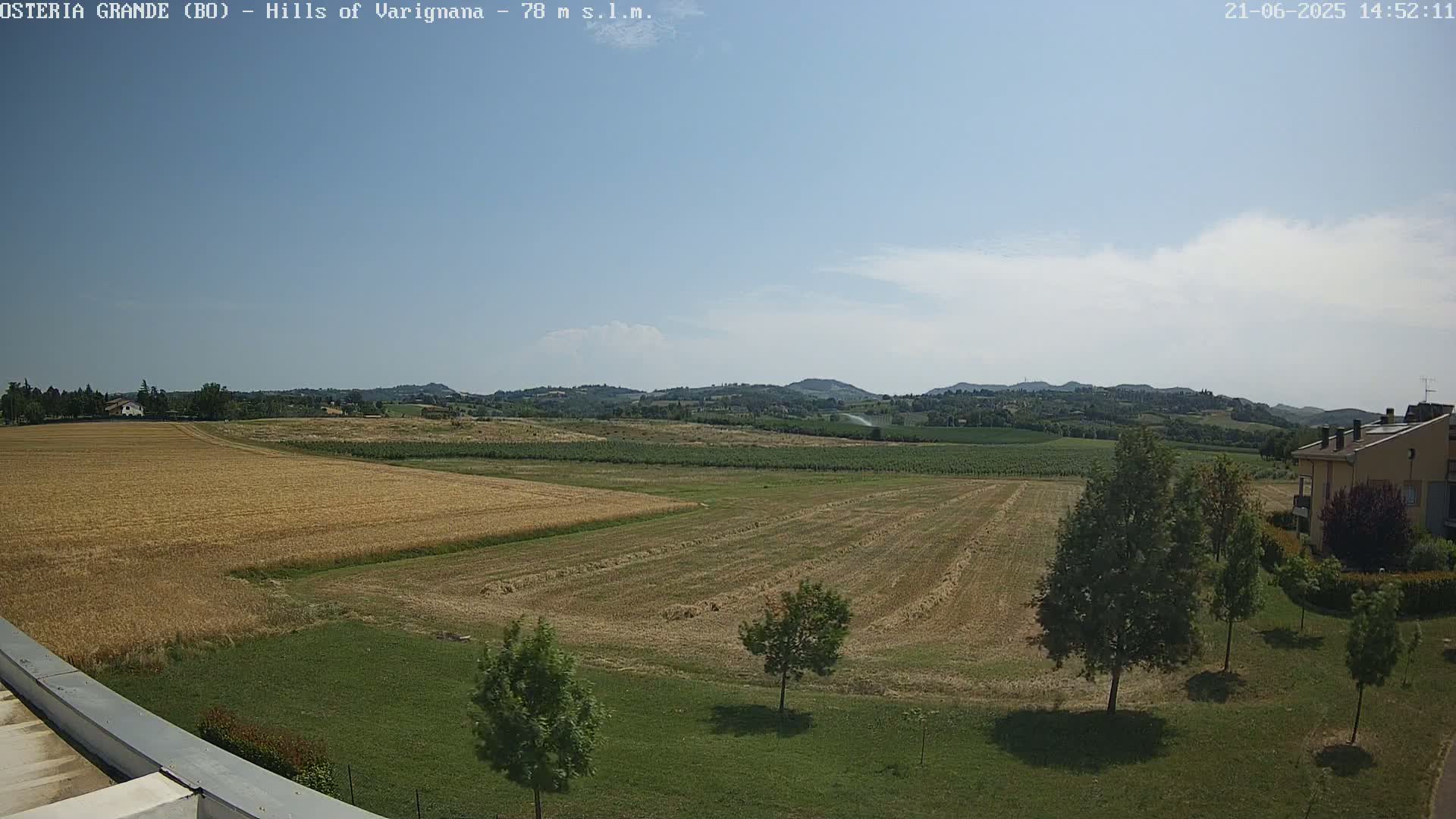 A mostly sunny day reveals a flat, harvested field bordered by green grass, a few trees, and distant rolling hills under a light blue sky.