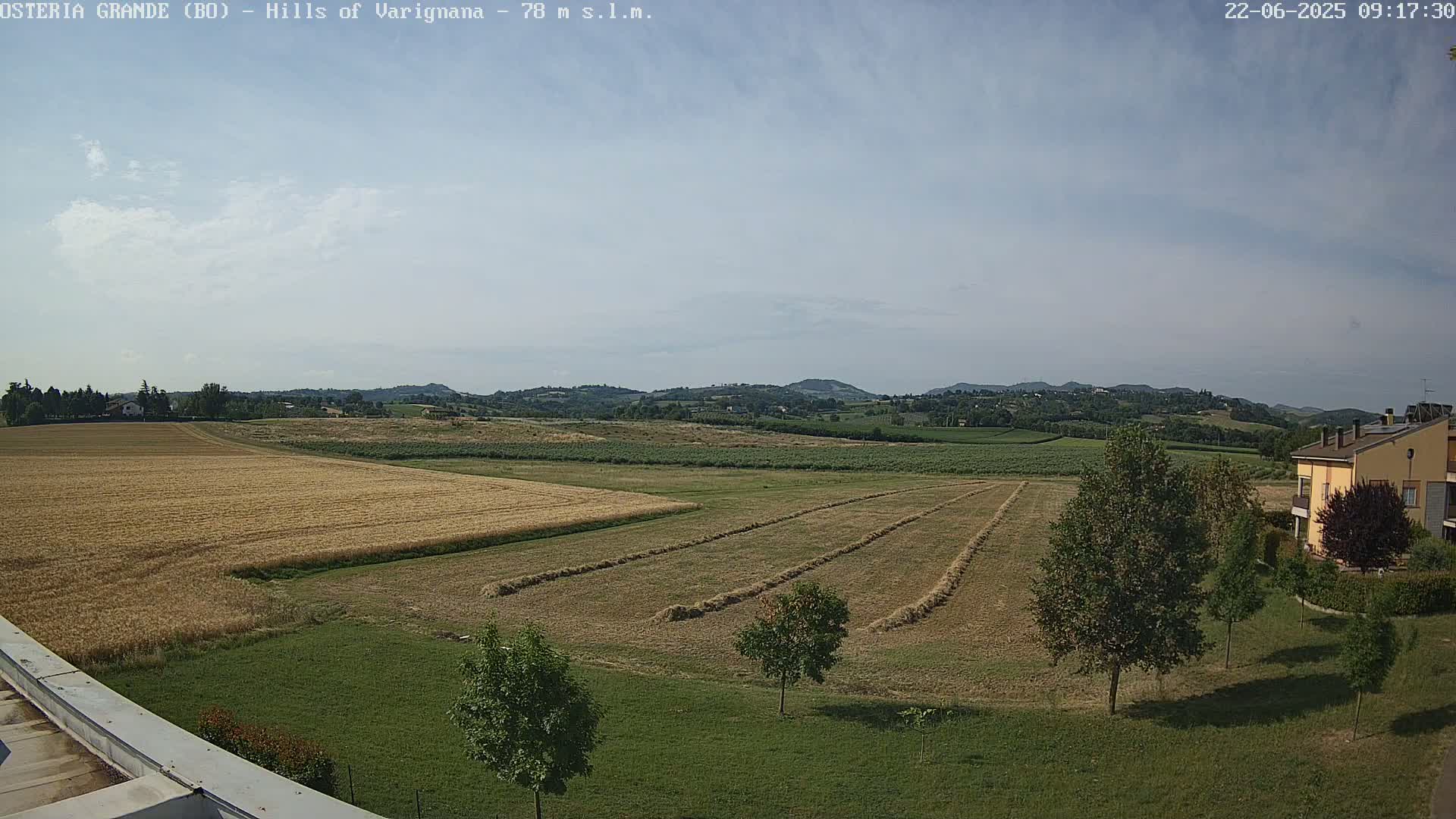 A partly sunny day reveals a rural landscape of harvested and unharvested fields, with rows of hay bales in the foreground and distant hills under a pale sky.