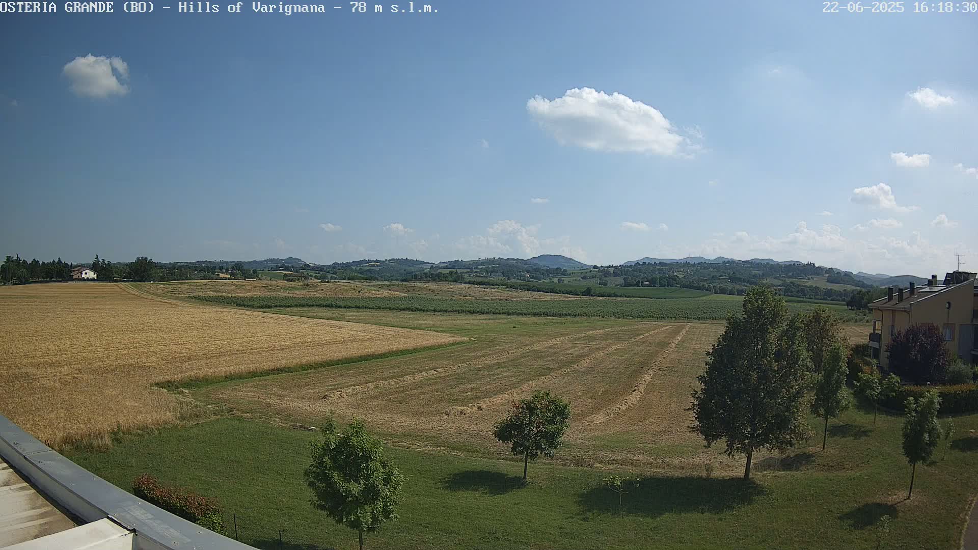 A partly sunny day reveals a harvested field, a few houses, and rolling hills in the distance.