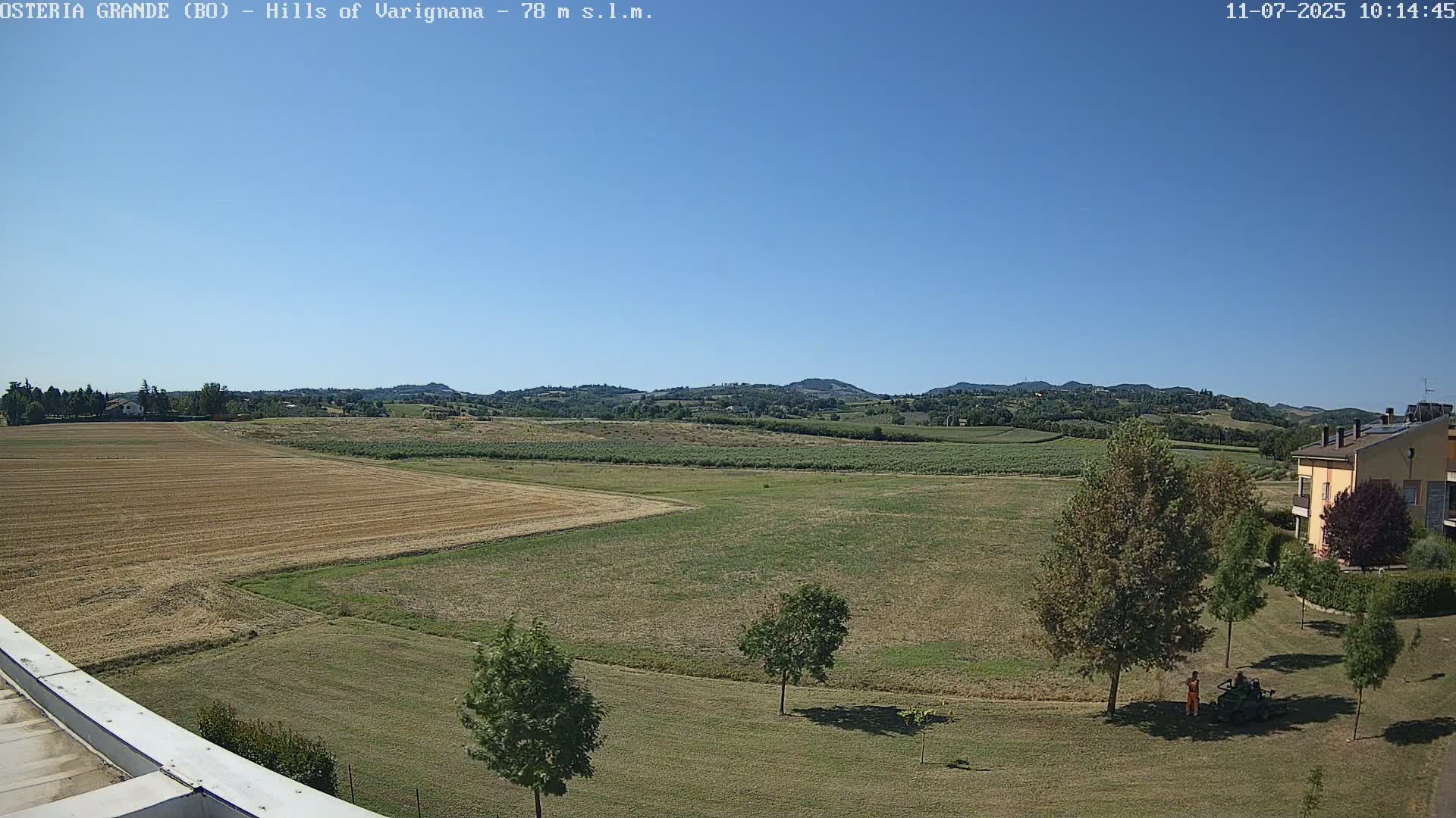 A wide, sunny view of rolling hills and farmland, with a few buildings visible in the distance and two people operating a piece of lawn equipment in the foreground.