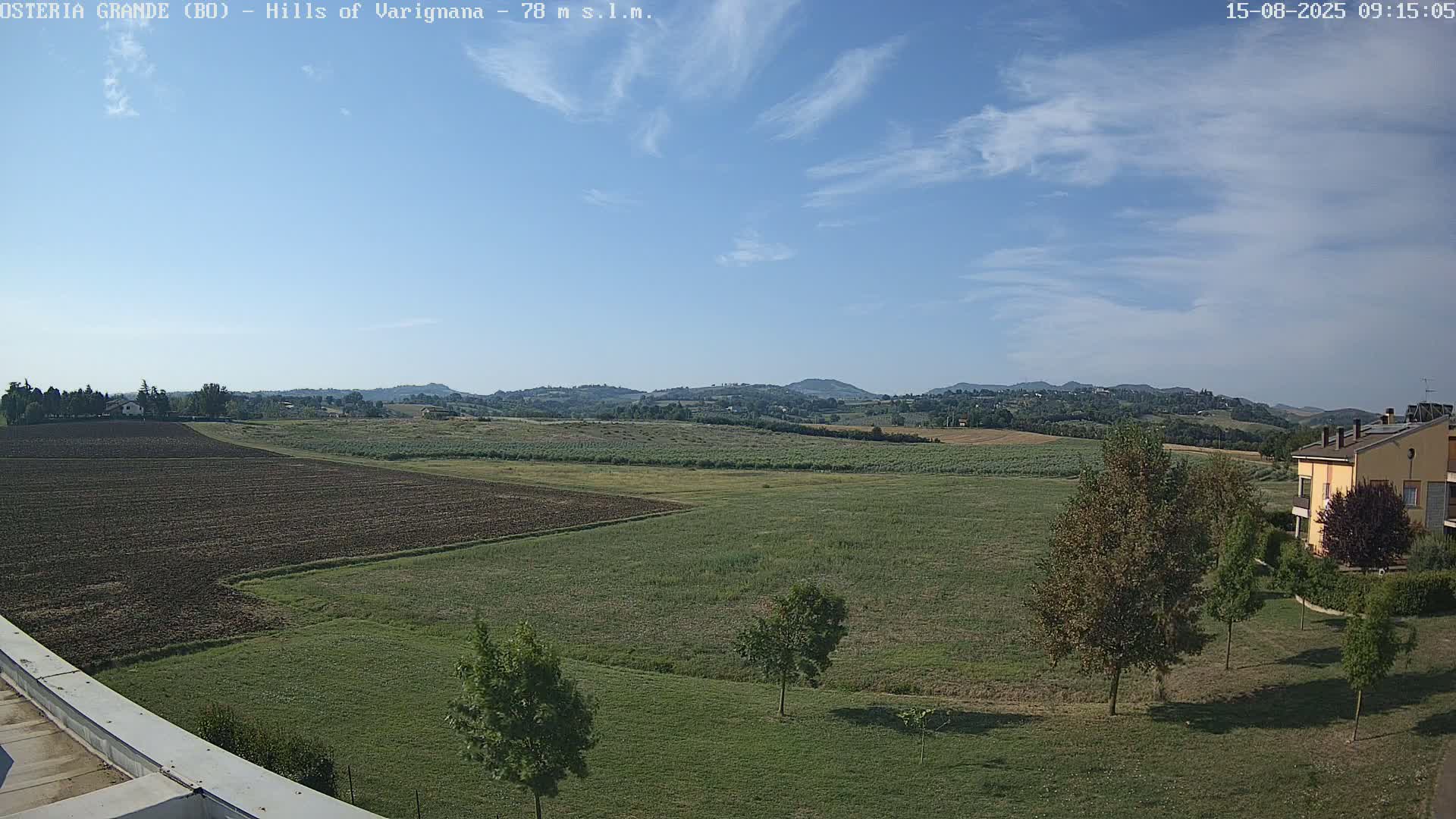 A partly cloudy sky overlooks a flat expanse of farmland, punctuated by a few trees and a building in the lower right, with rolling hills visible in the distance.