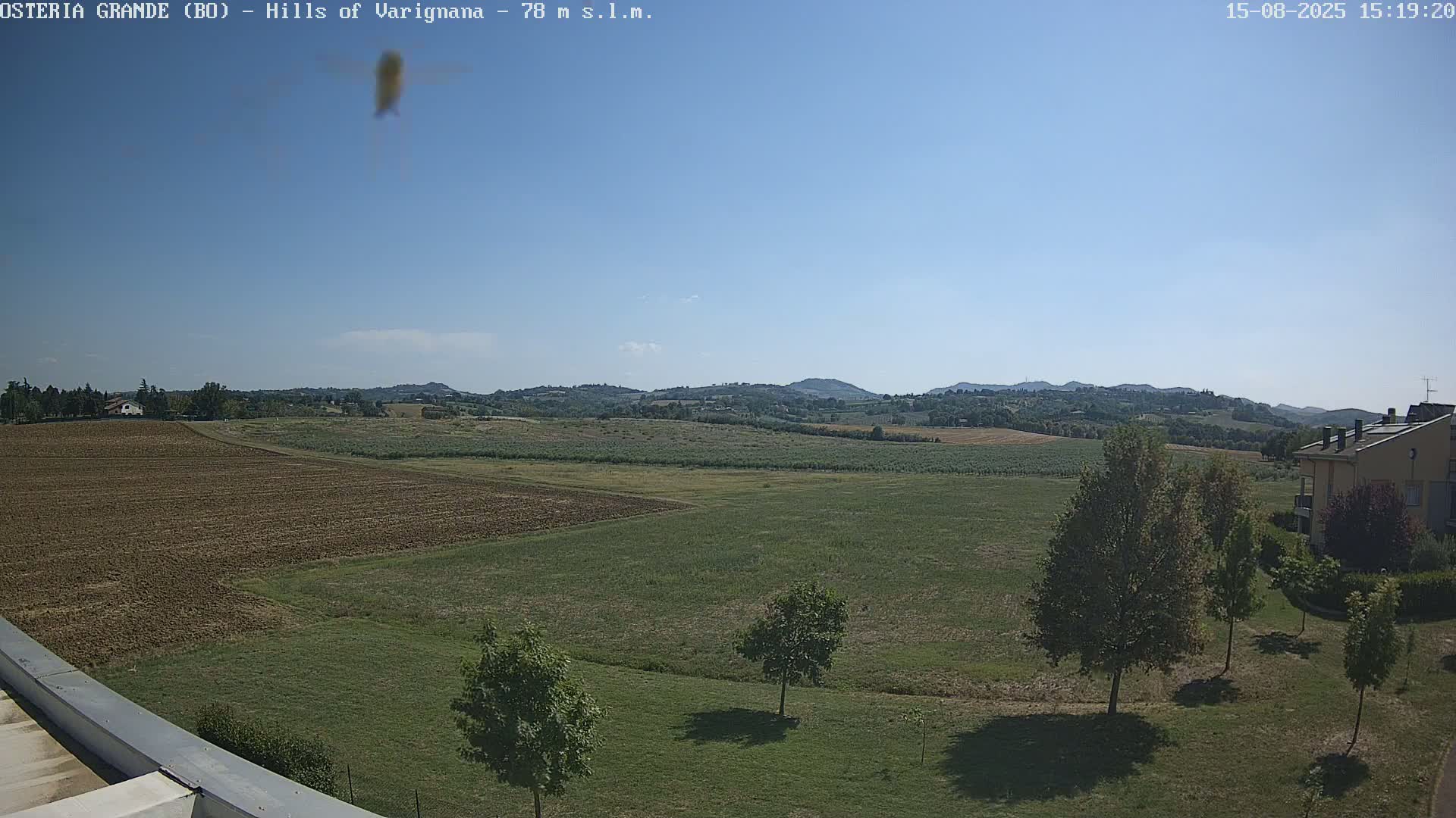 A partly cloudy sky overlooks a flat expanse of farmland, punctuated by a few trees and a building in the lower right, with rolling hills visible in the distance.