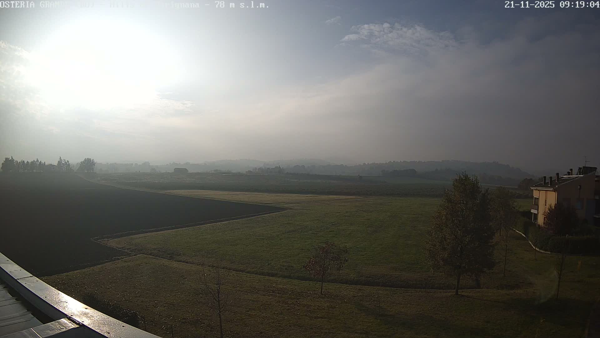 A hazy morning view captures extensive agricultural fields and rolling hills receding into the misty distance, beneath a bright, overcast sky with the sun shining through, alongside some trees and a building on the right.