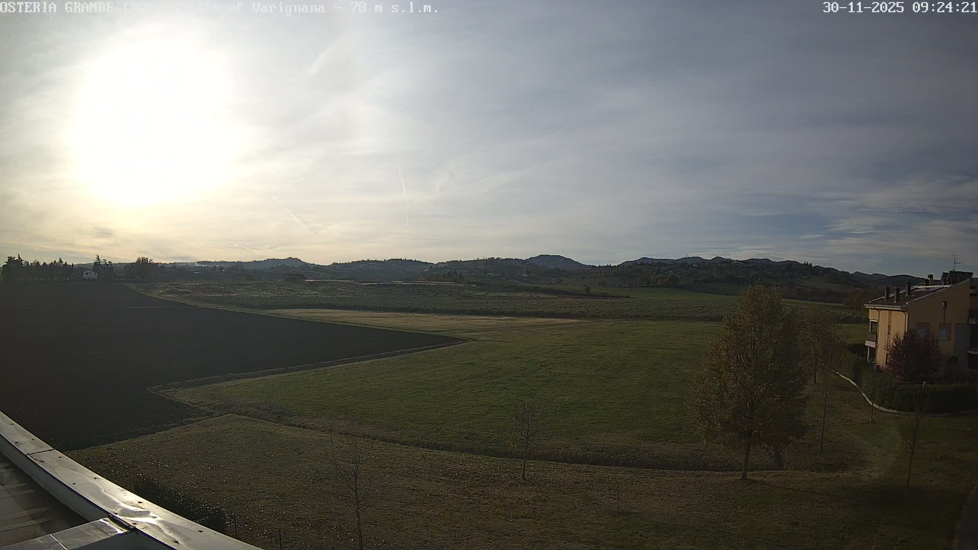 Under a bright, partly cloudy sky with a prominent sun, a vast rural landscape features green and tilled fields extending towards distant forested hills, with a multi-story building and trees with autumn foliage on the right.