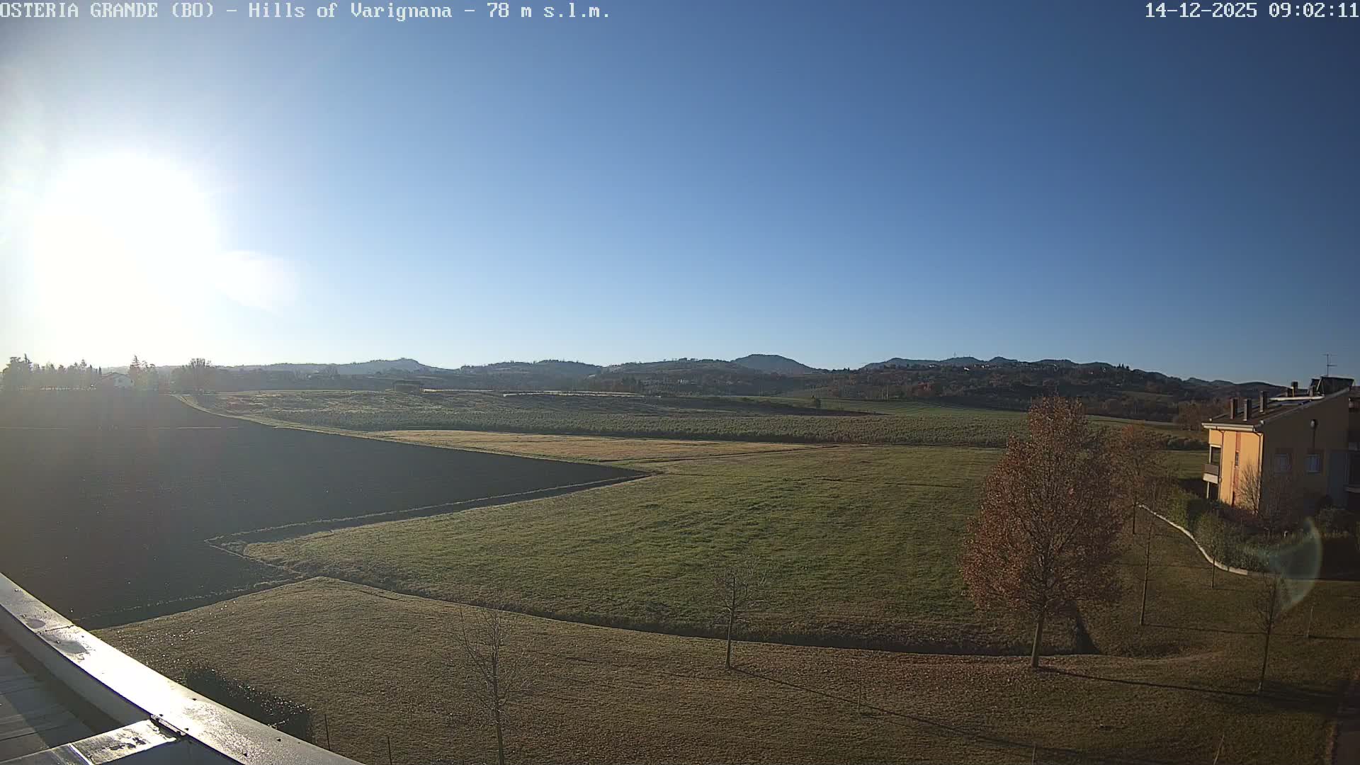 A bright sunny day with a clear blue sky illuminates a landscape of rolling hills and agricultural fields, with a building and a leafless tree partially visible in the foreground.