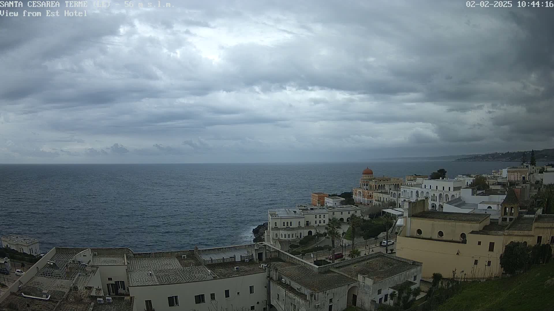 A coastal town with white buildings sits on a cliff overlooking a dark blue sea under a cloudy sky.