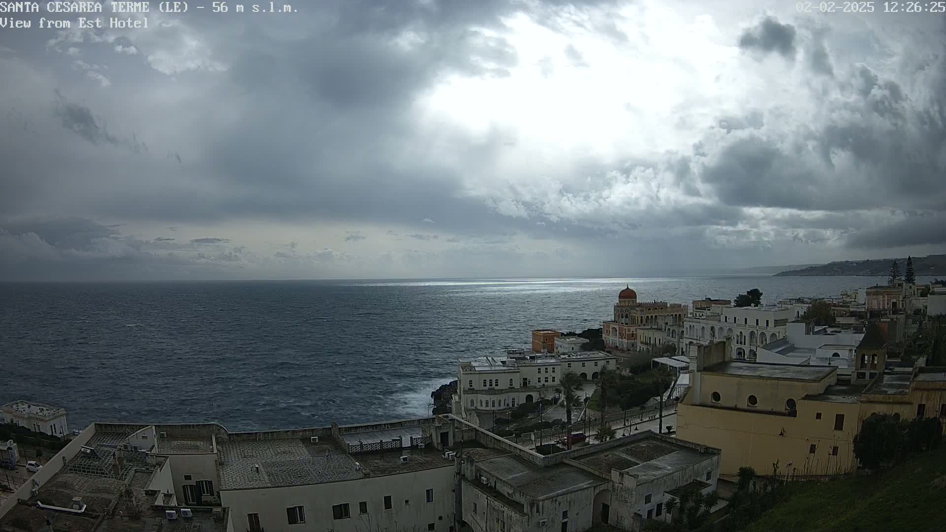 A coastal town with white buildings under a cloudy sky overlooks a dark, choppy sea.