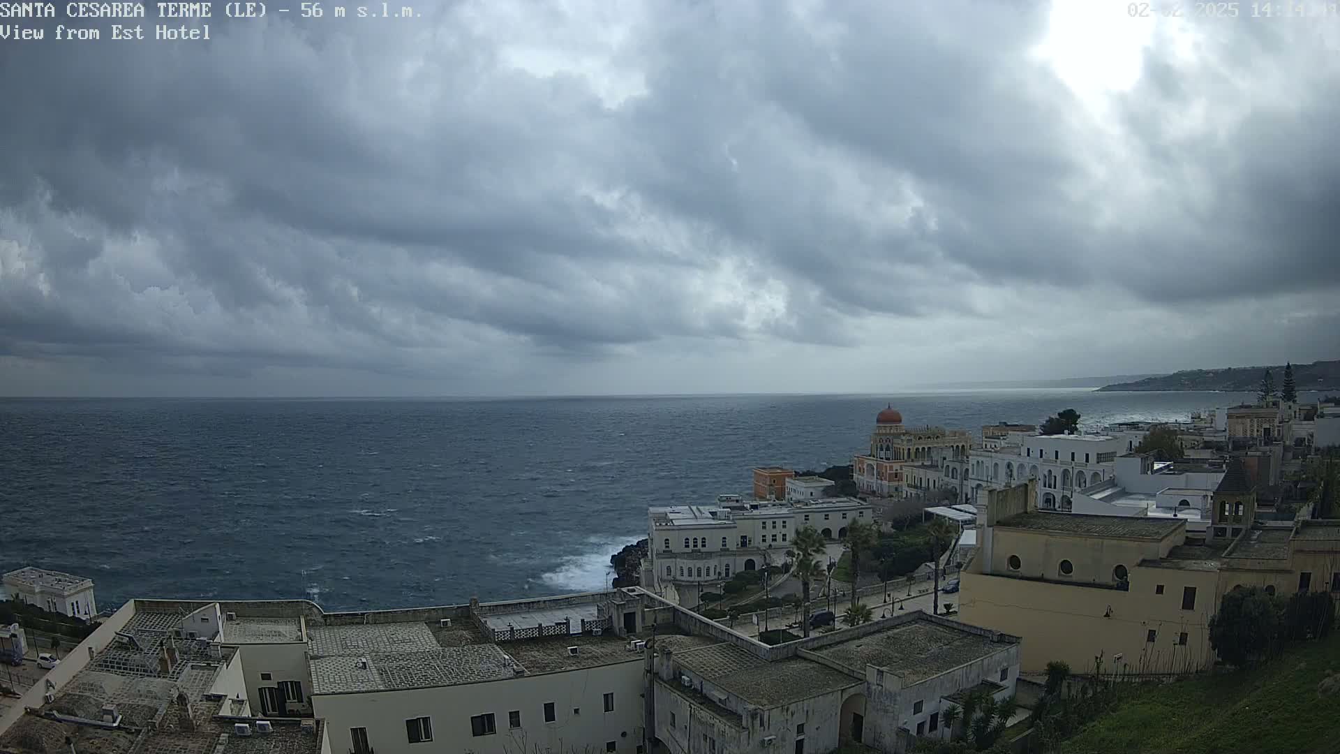 A coastal town with white buildings sits on a cliff overlooking a choppy sea under a cloudy sky.