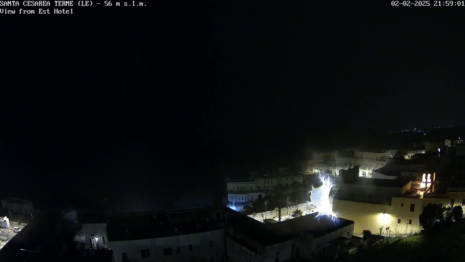 A nighttime view of a town, possibly coastal, with fog or mist obscuring the lower parts of the scene under a dark sky, illuminated by streetlights and building lights.