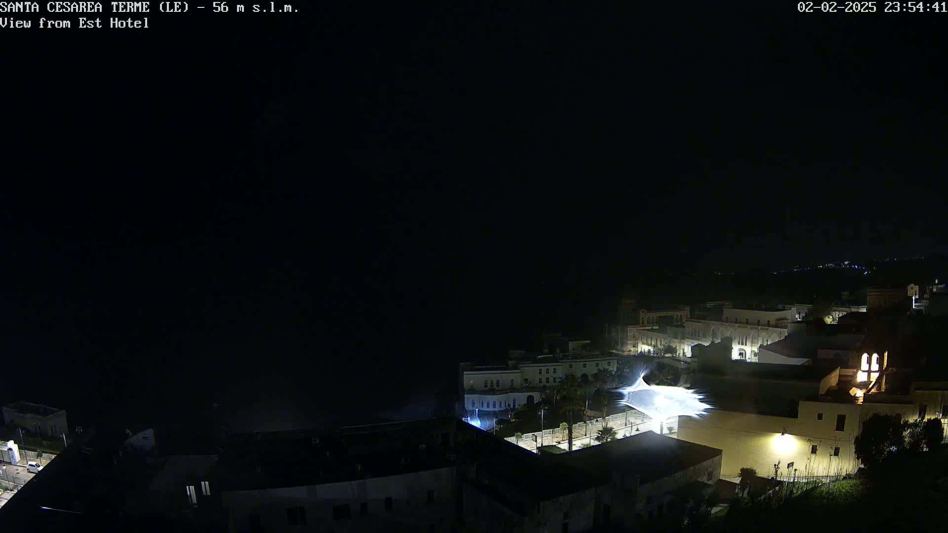 A nighttime, coastal town is illuminated, with a bright light source emanating from a central point in the lower middle section of the image.