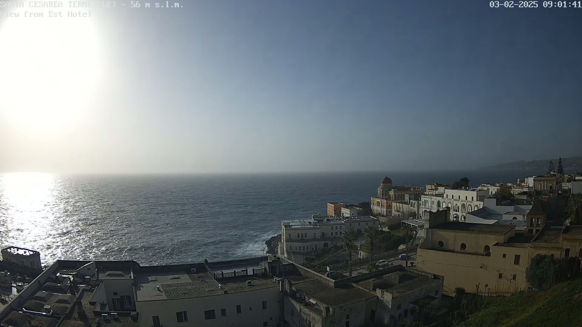 A hazy sun shines over a calm sea, overlooking a coastal town of white buildings with a few red-roofed structures nestled against a cliffside.