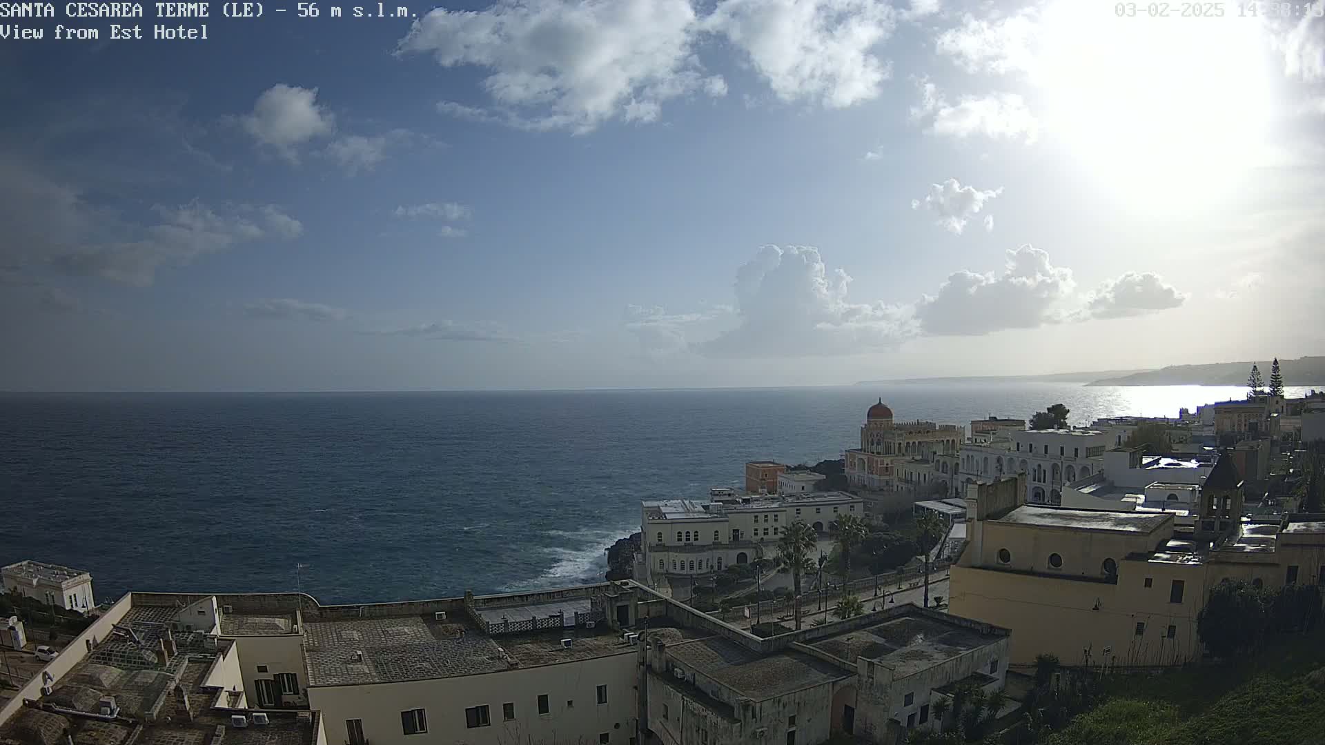 A coastal town with white buildings and a red-domed structure sits on a cliff overlooking a dark blue sea under a partly sunny sky with fluffy white clouds.