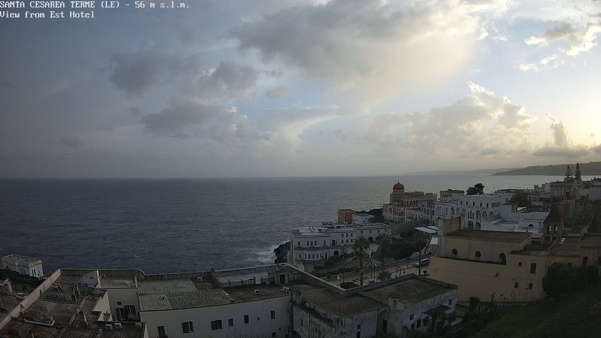 A coastal town with white buildings sits on a rocky shoreline overlooking a dark blue sea under a cloudy sky.