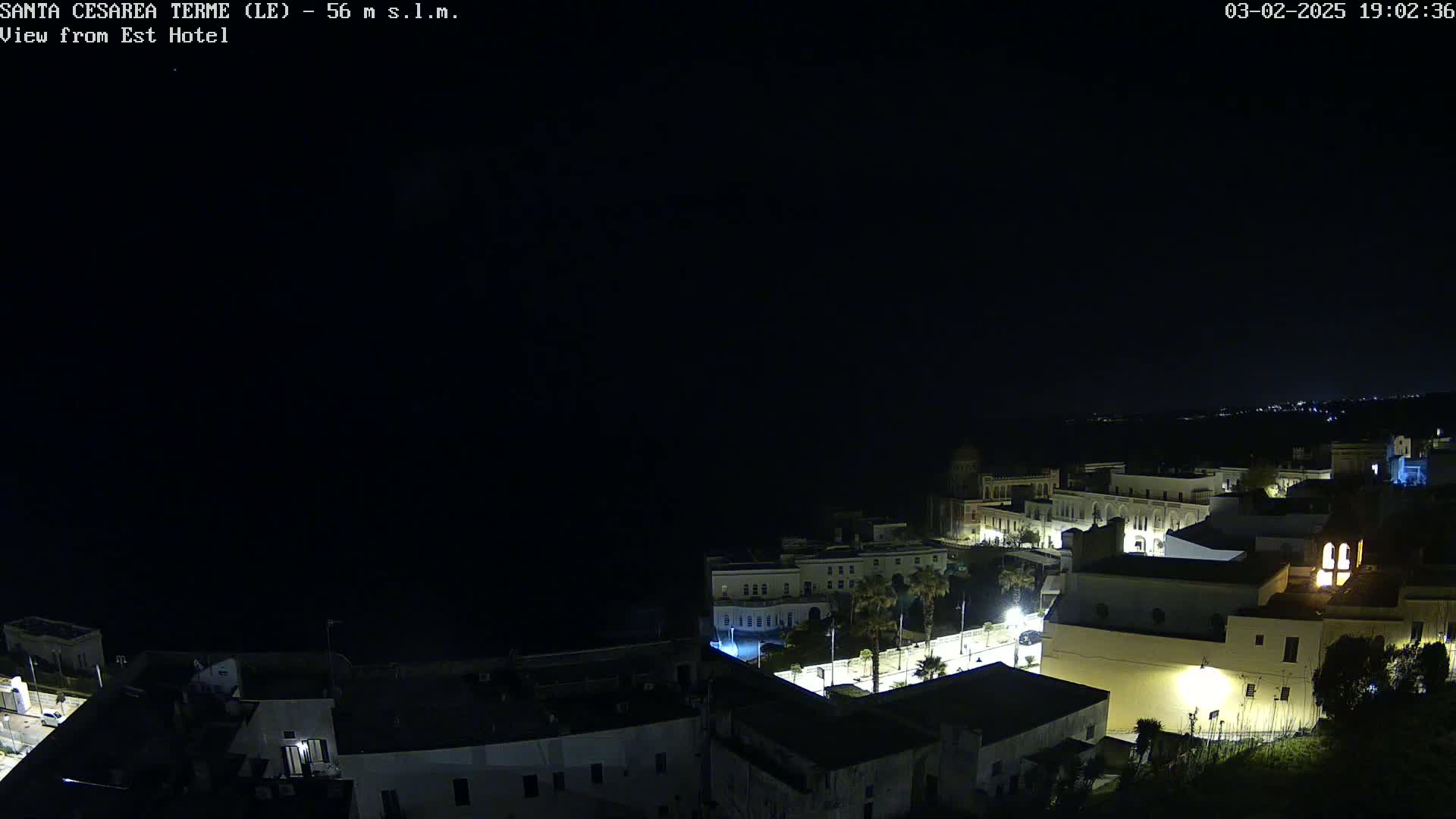 A nighttime, aerial view of a coastal town shows dimly lit buildings near a dark body of water under a clear, dark sky.