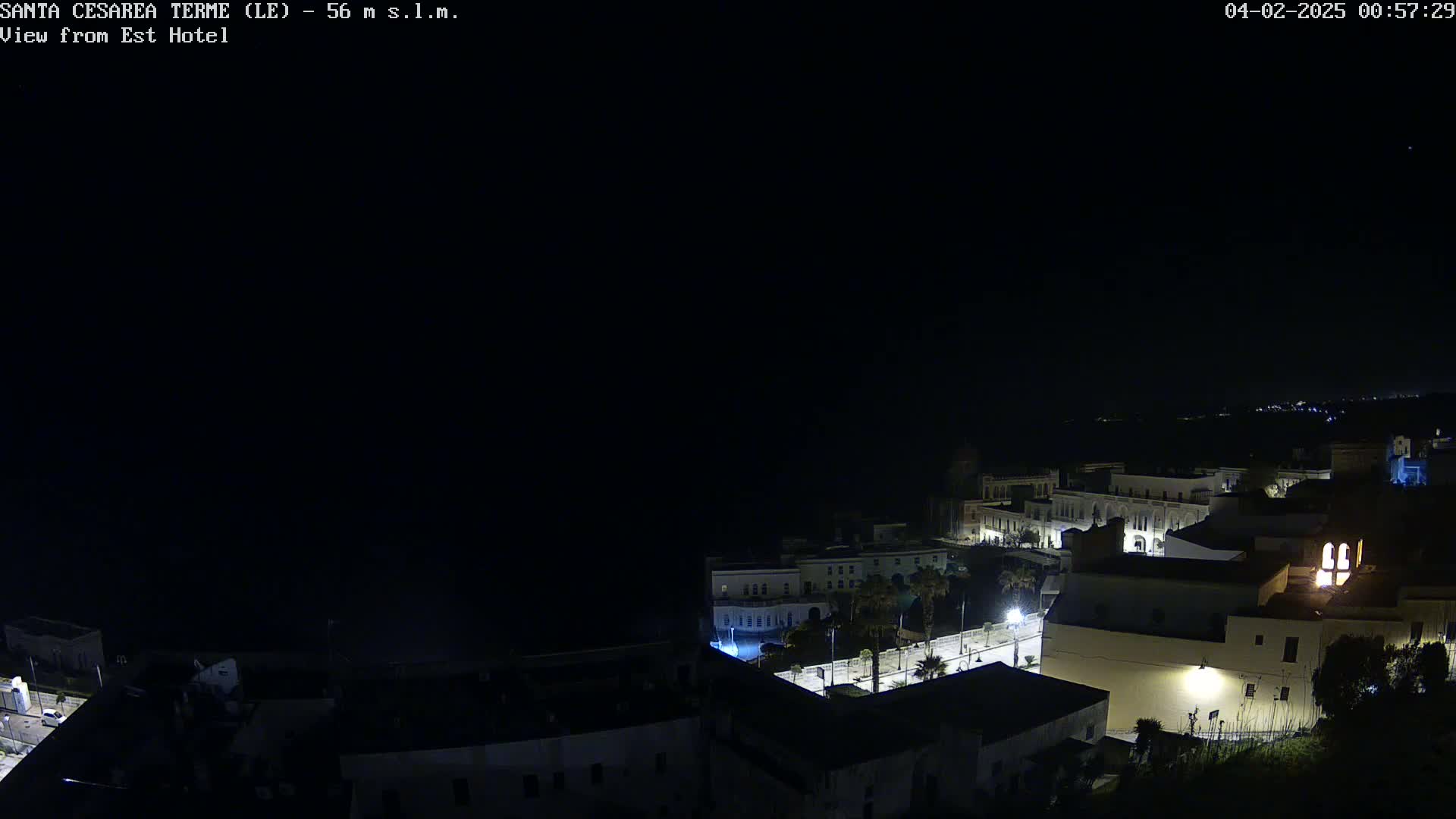 A nighttime aerial view of a coastal town shows dimly lit buildings near a dark ocean under a clear, starlit sky.